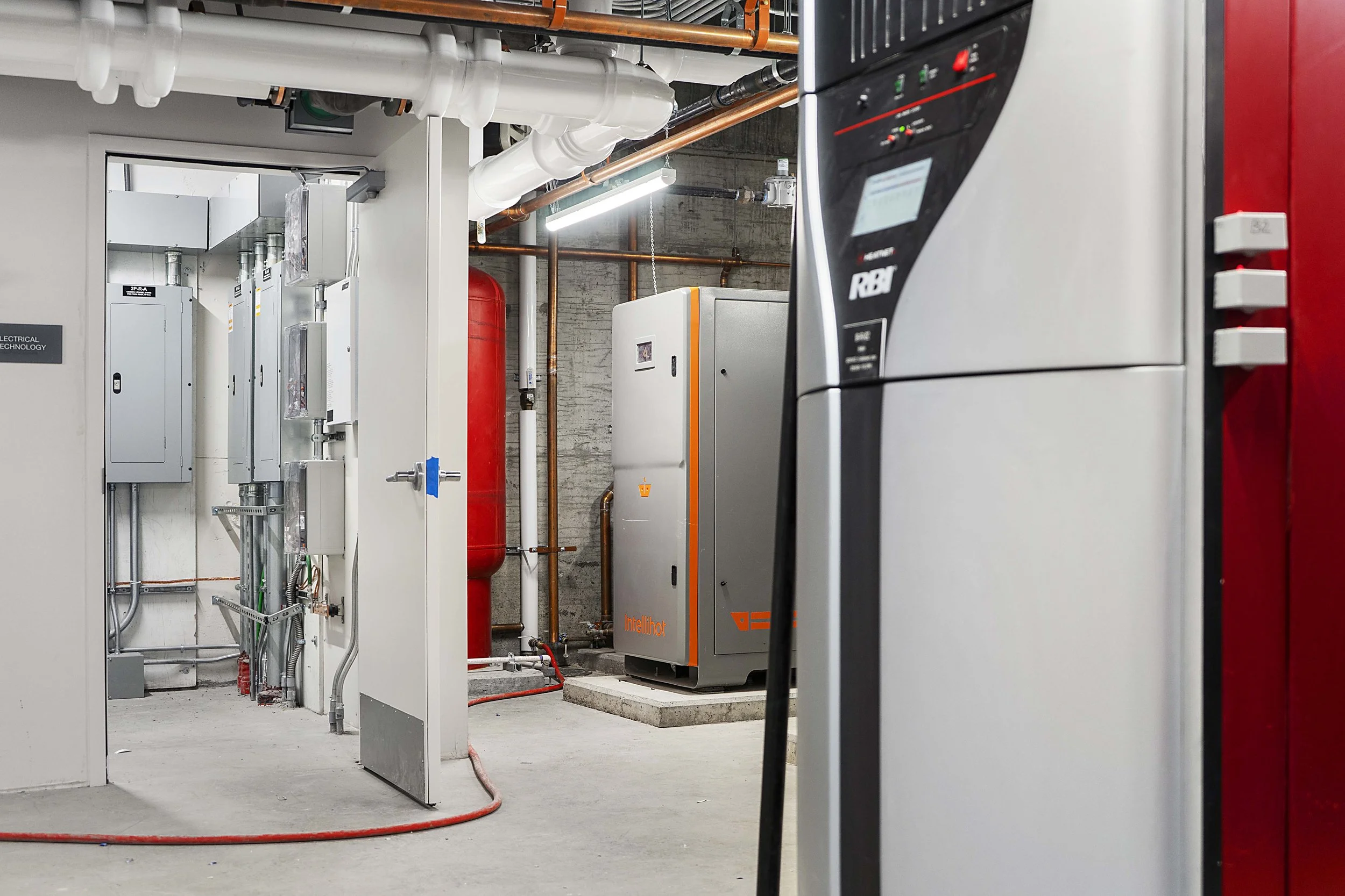 A room with industrial HVAC and electrical equipment, including a large metal control panel on the right, smaller electrical boxes on the left, and piping and red tanks in the background.