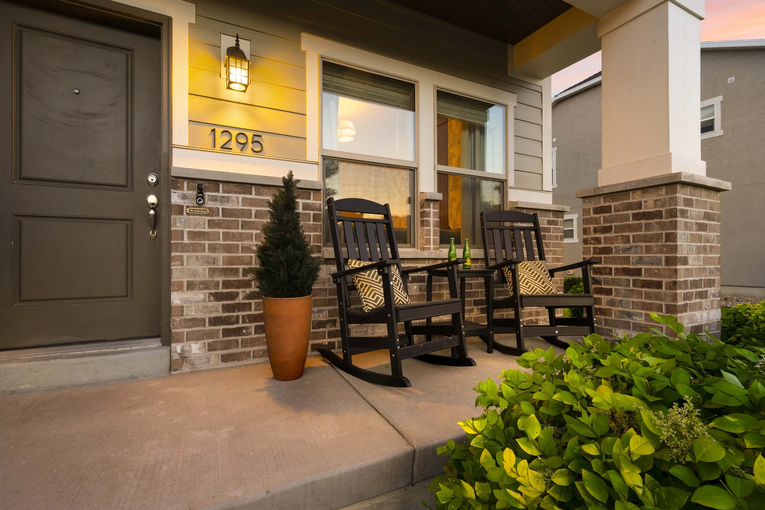 The front porch of a house with two black rocking chairs, green bottles on the table, a potted plant, and a yellow house number 1295, illuminated by a porch light at sunset.