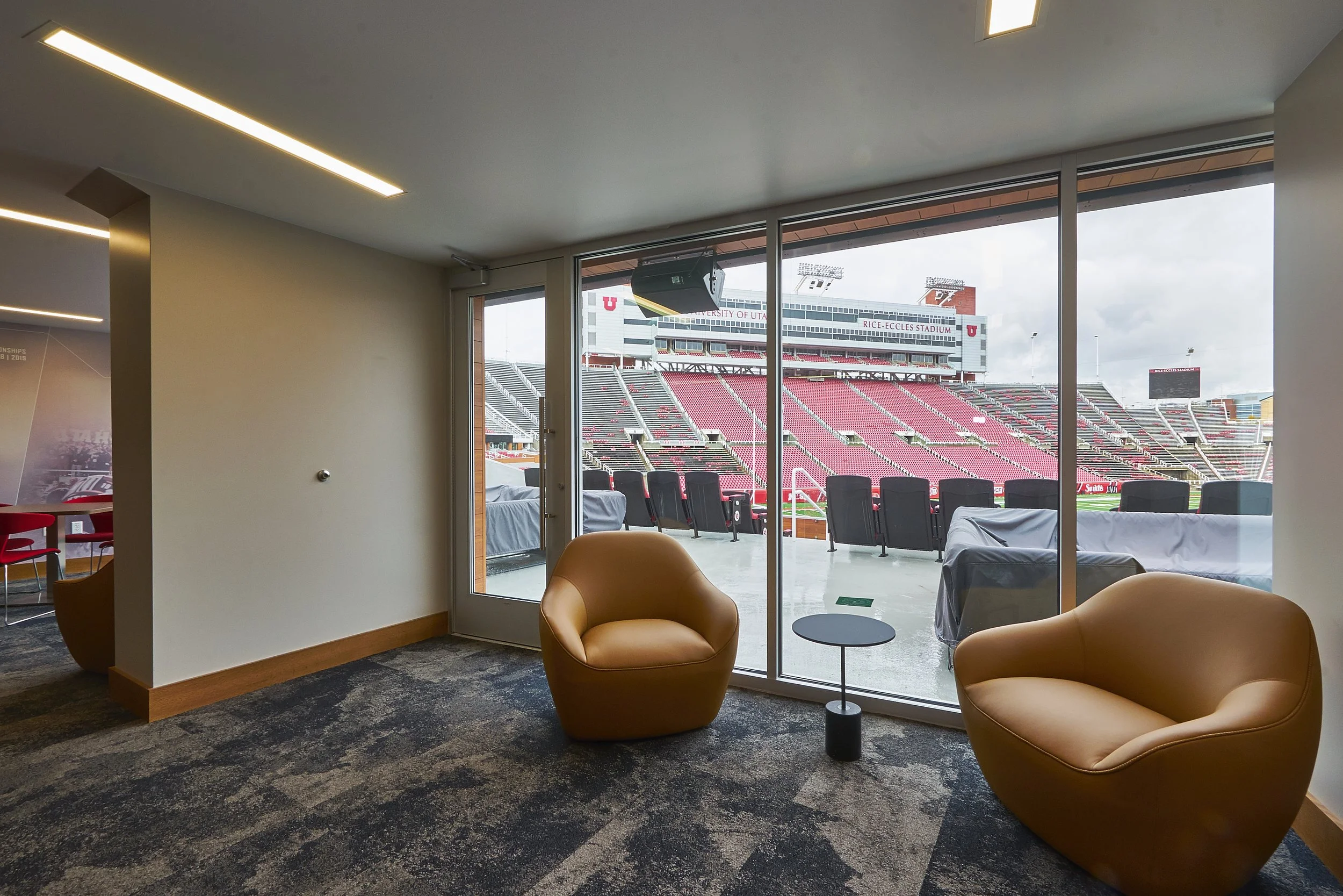 View of the Utah Utes football stadium from inside a lounge area with two tan armchairs, a small side table, and large glass doors opening to the stadium seating outside.
