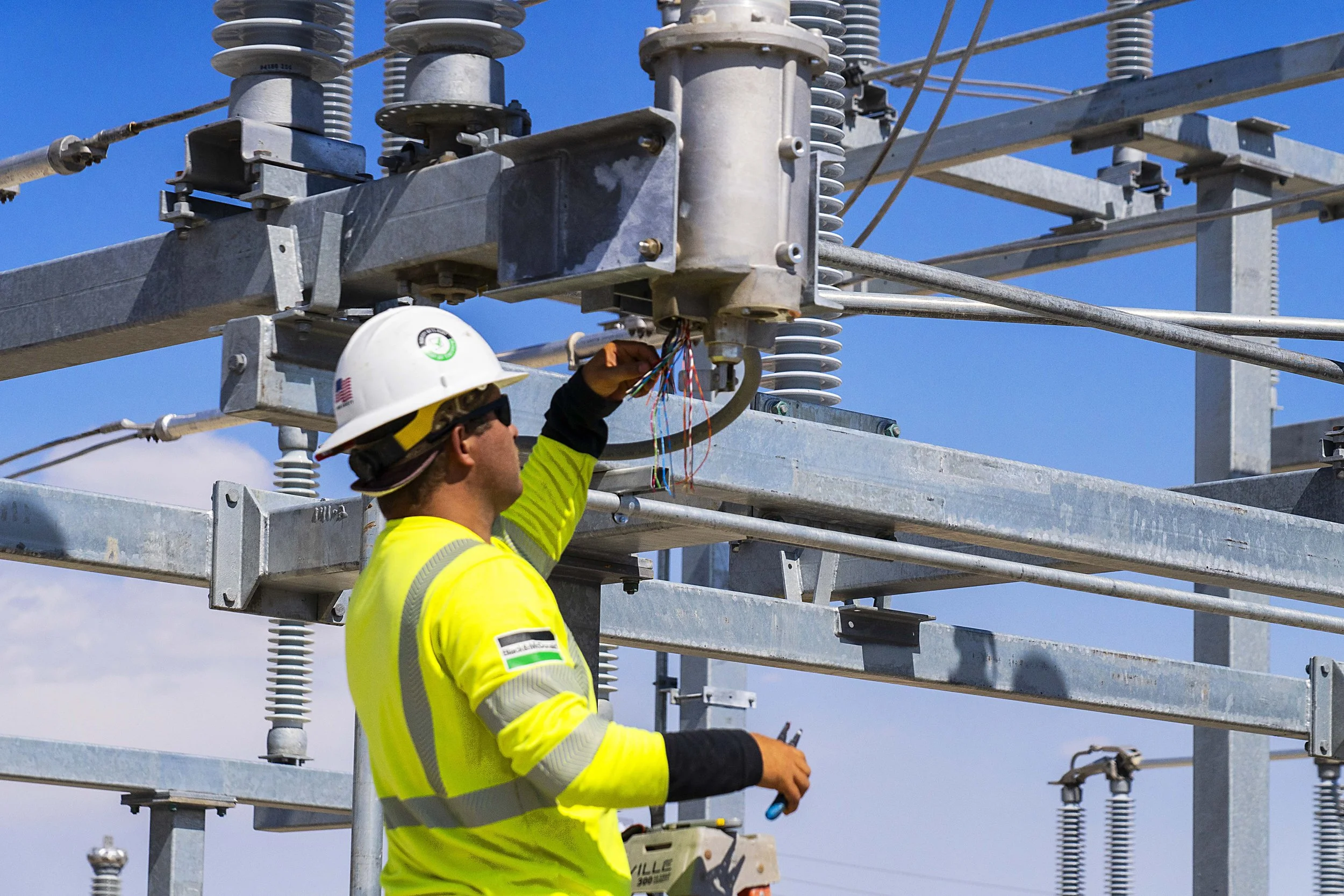Worker in yellow safety gear and white helmet working on electrical components of a power transmission tower under a blue sky.