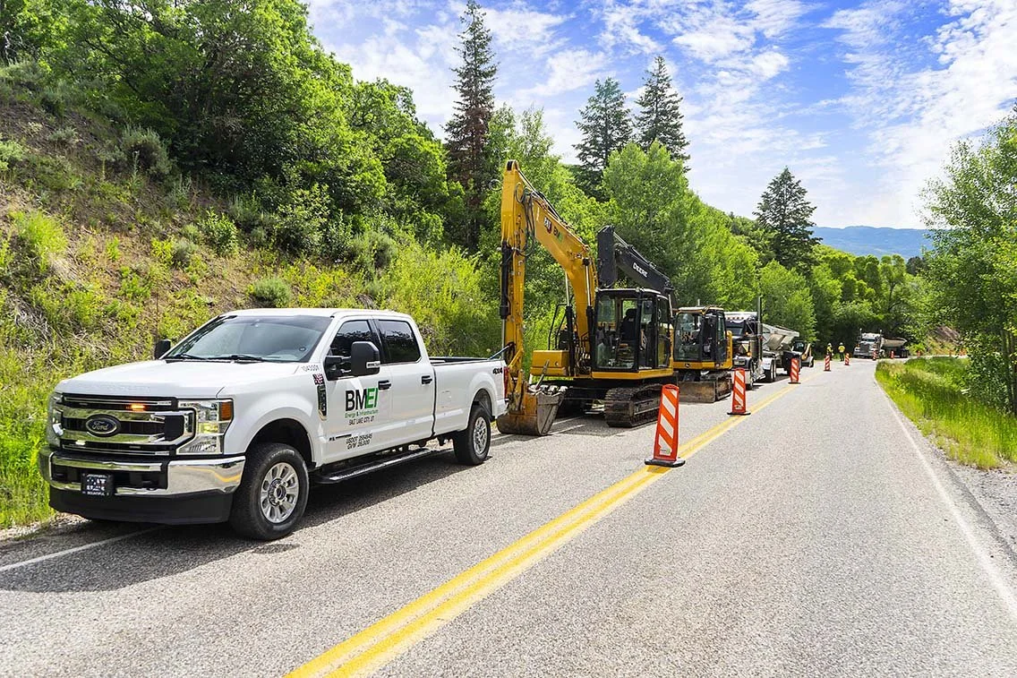 Construction site, White Ford Company truck with multiple track hoe style tractor's, single lane road with construction cones 