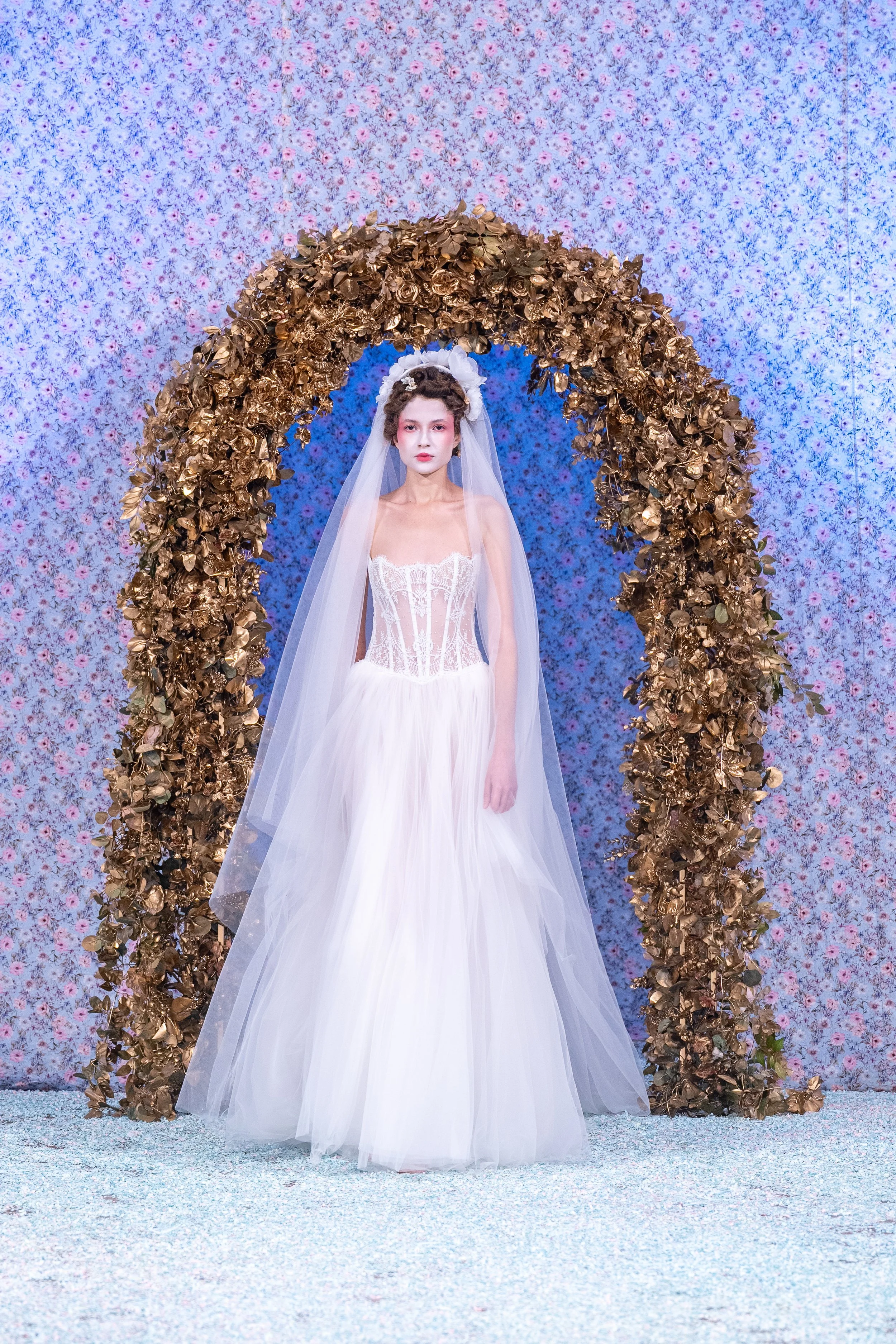 Model in a white wedding dress and veil standing under a gold floral arch, with a floral pink and blue backdrop.