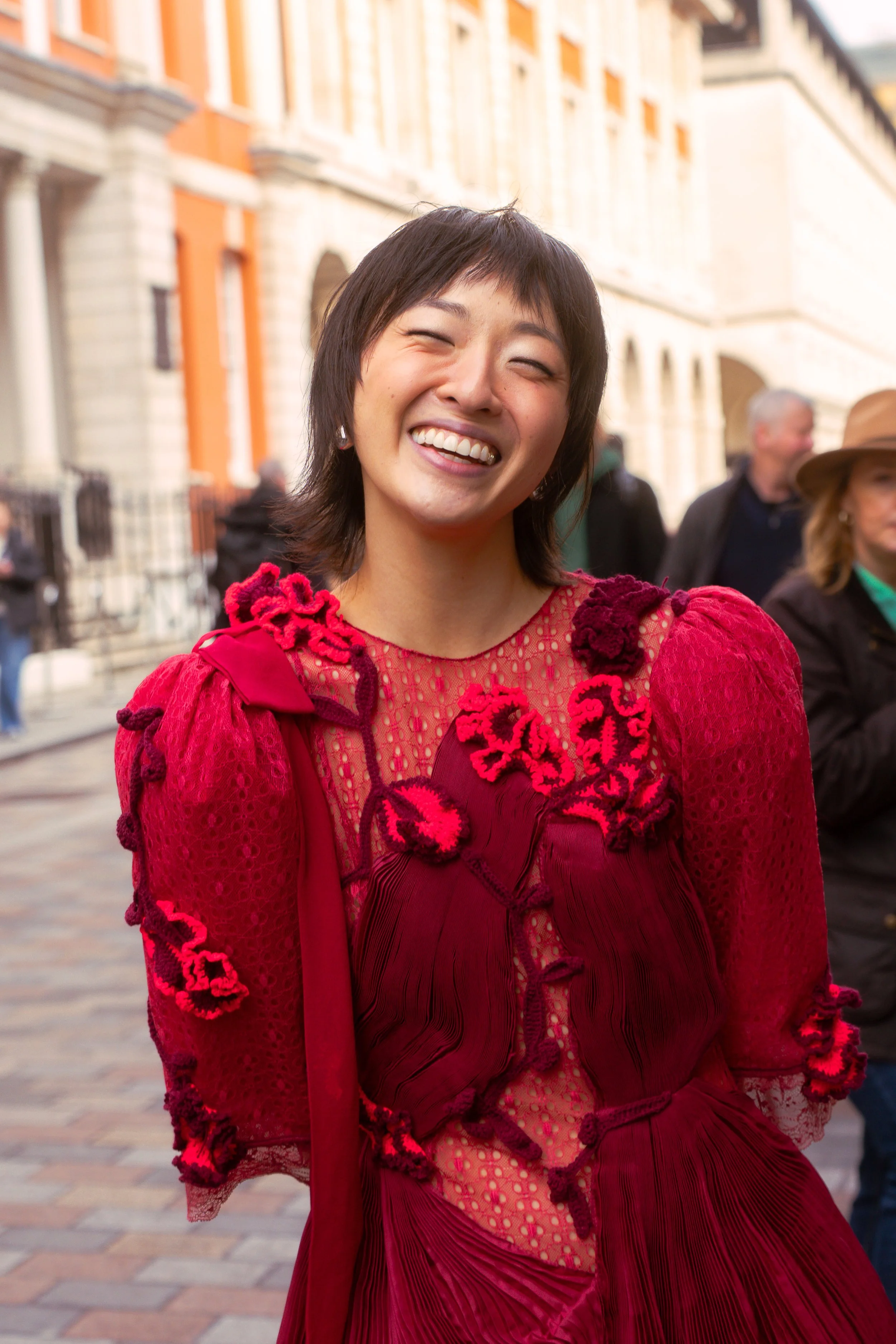 A woman with short black hair smiling, wearing a vibrant red dress with intricate floral and textured details, standing on a city street with historic buildings and other people in the background.