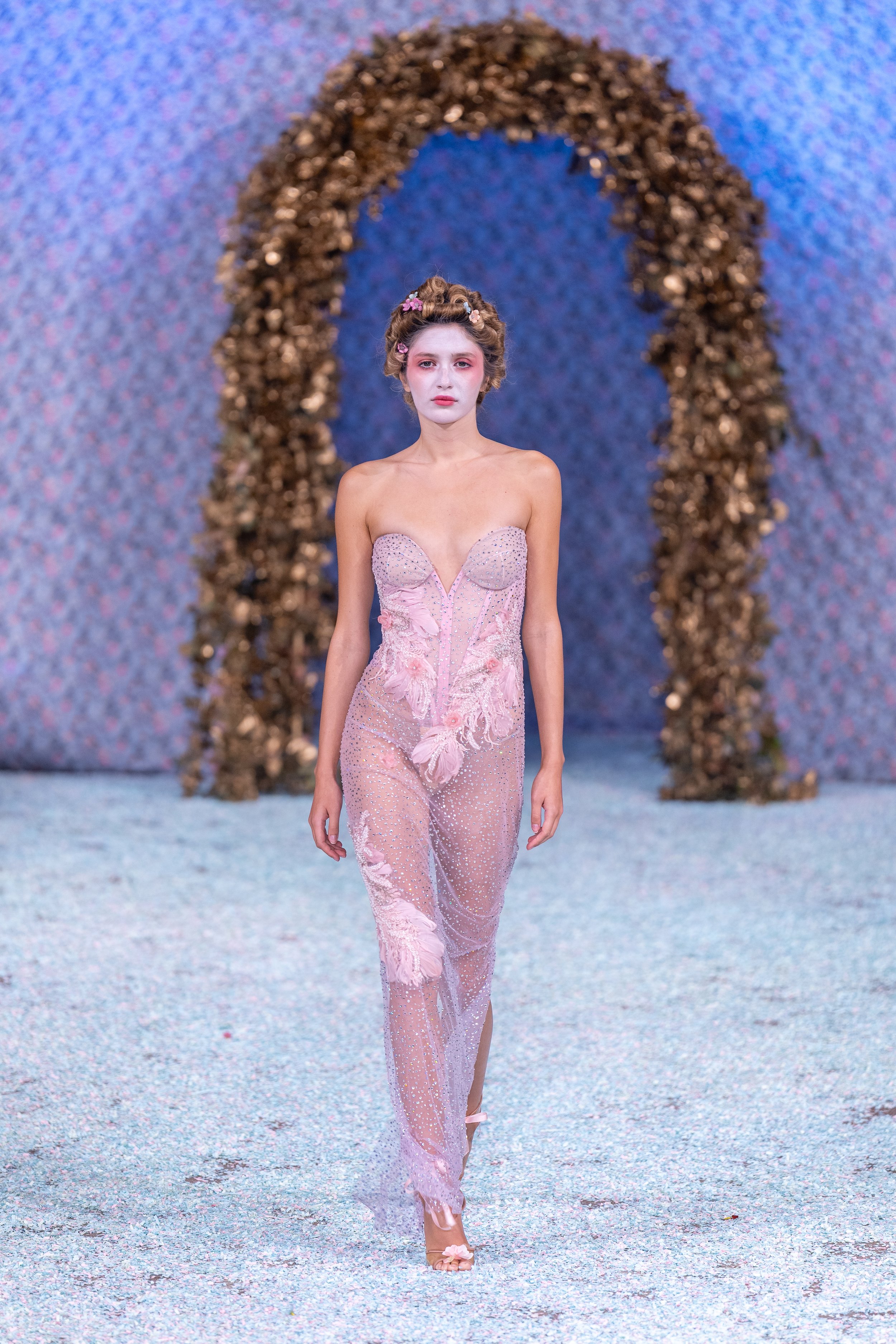 A model walking on a runway with a floral backdrop and arch, wearing a pink, sheer, embellished gown with floral details and pink flowers in her hair.