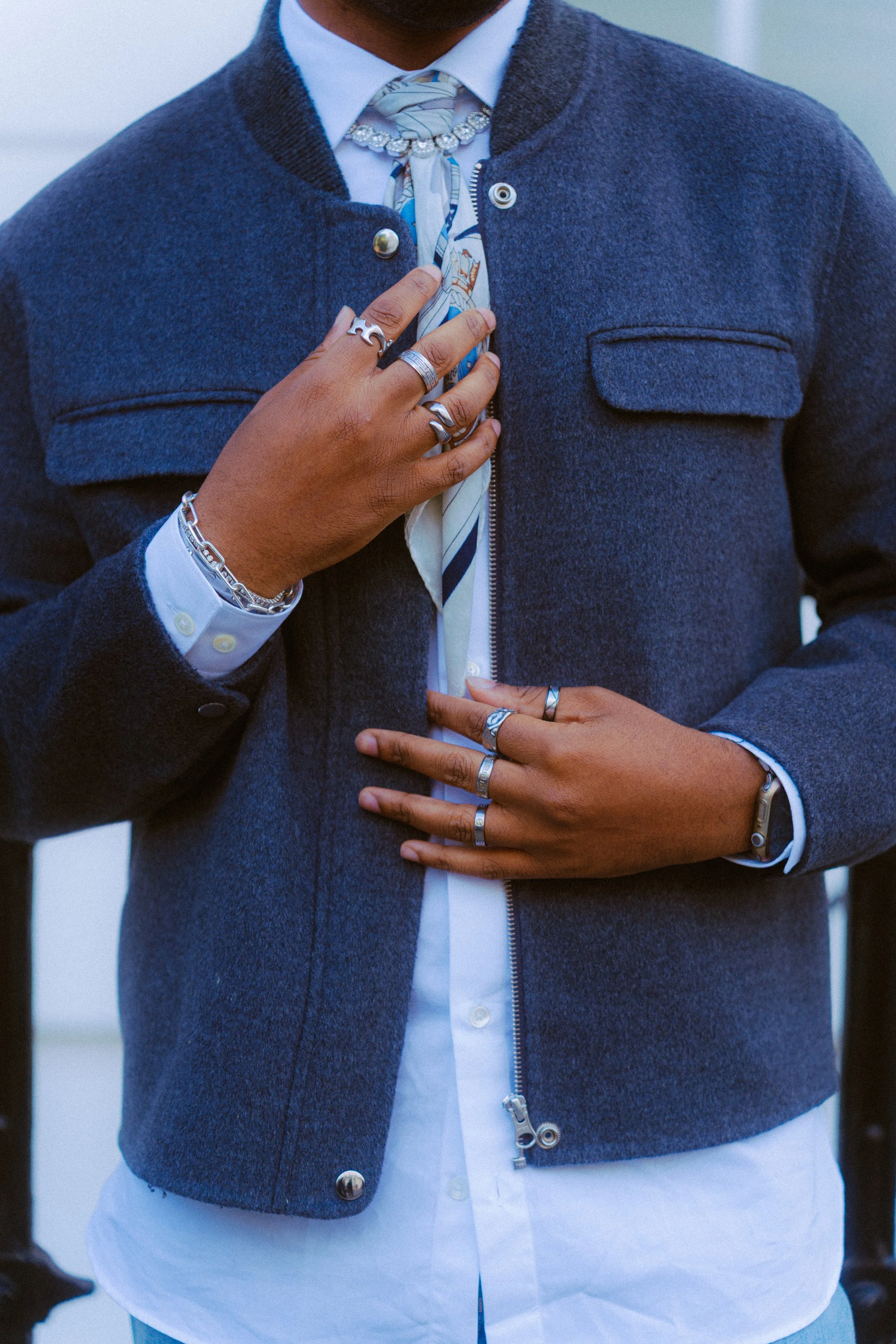 Close-up of a person's torso, wearing a navy blue jacket over a white shirt and accessorized with multiple rings, bracelets, and a watch.
