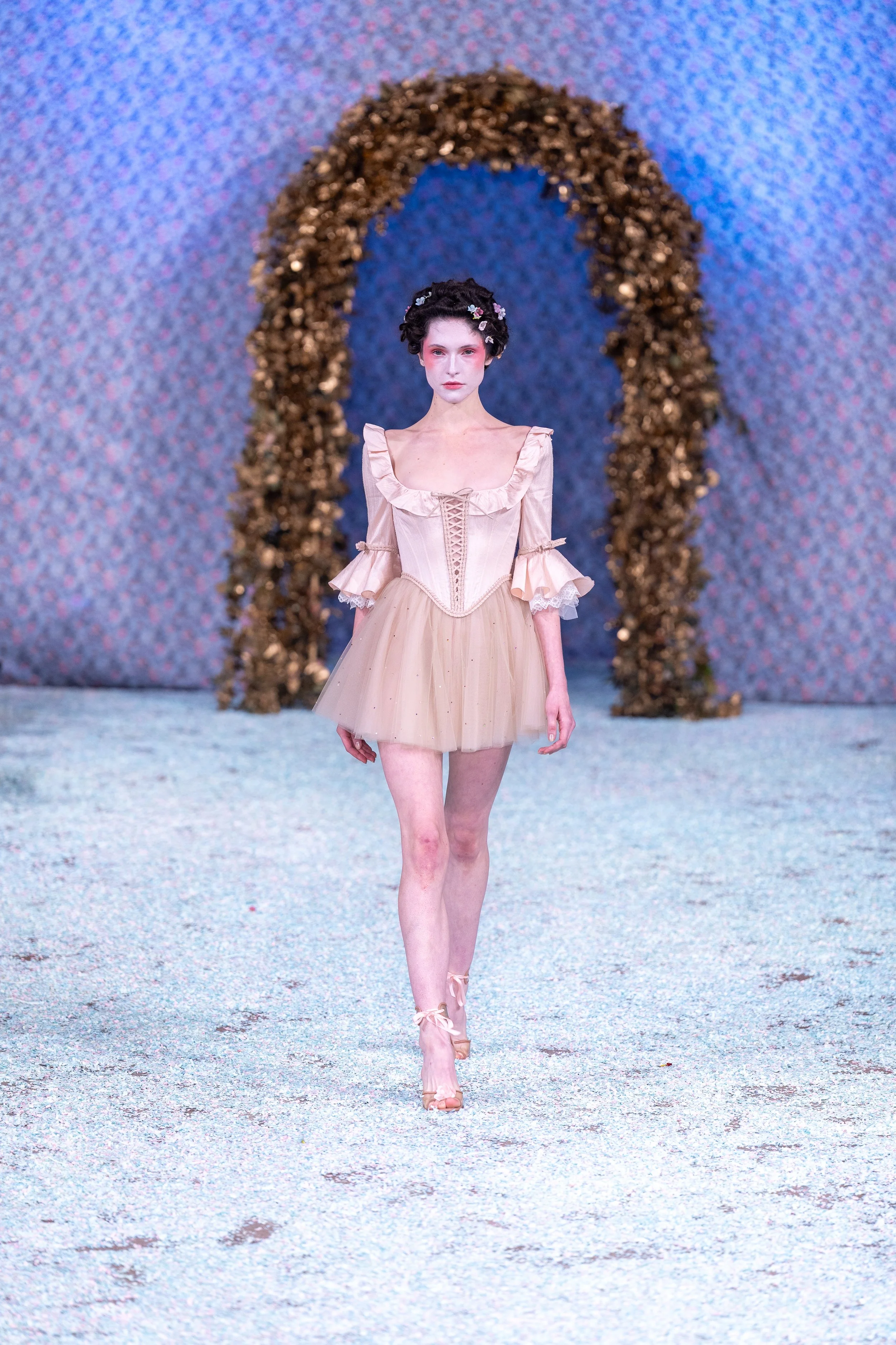 A model walking on a runway with a floral arch in the background, wearing a beige, ruffled, short dress with a corset-like bodice, pink flower headband, and ballet shoes.