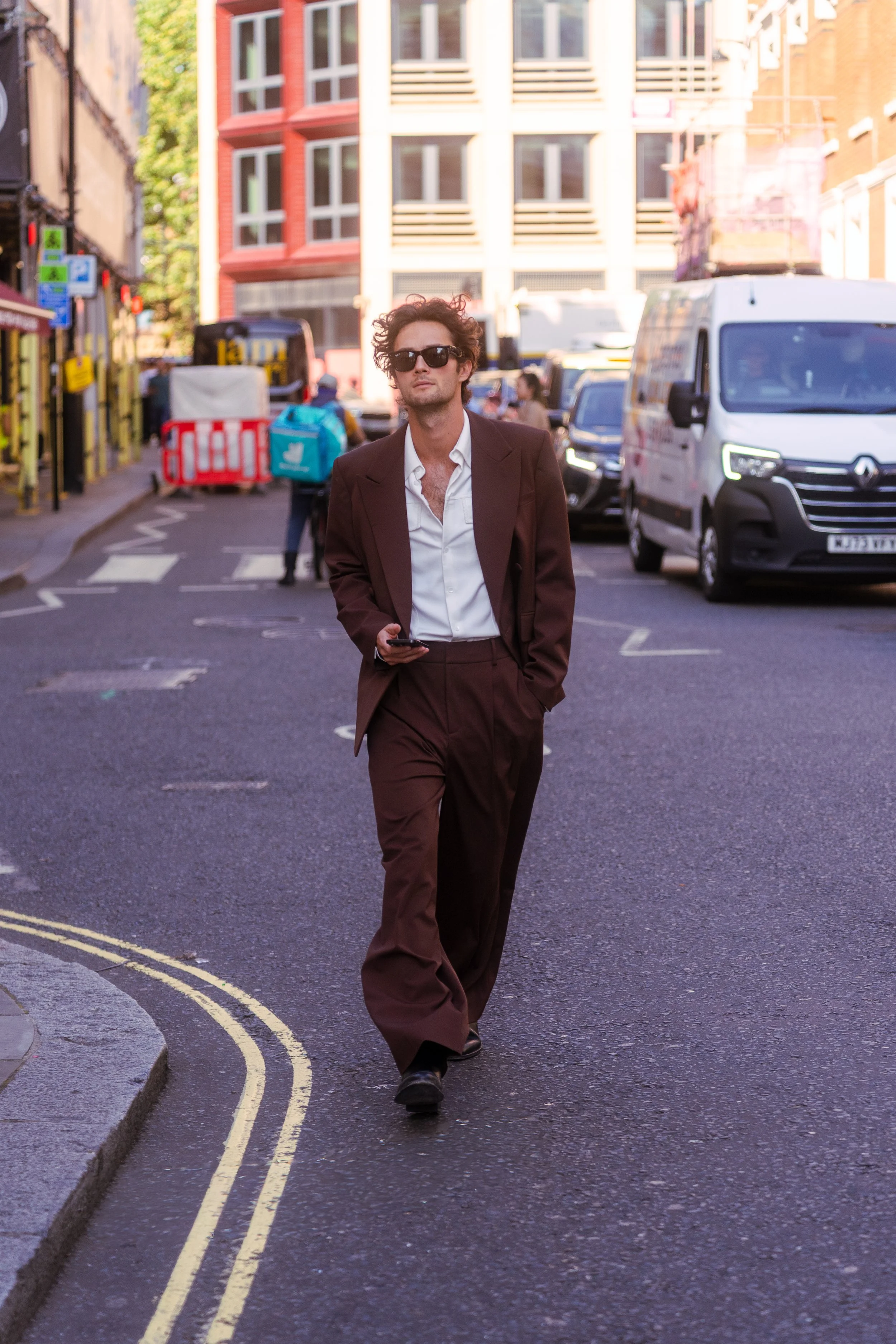 A man walking on a city street wearing sunglasses, a dark brown suit, and a white shirt, holding a smartphone in his right hand. There are parked cars and pedestrians in the background, along with colorful buildings and storefronts.