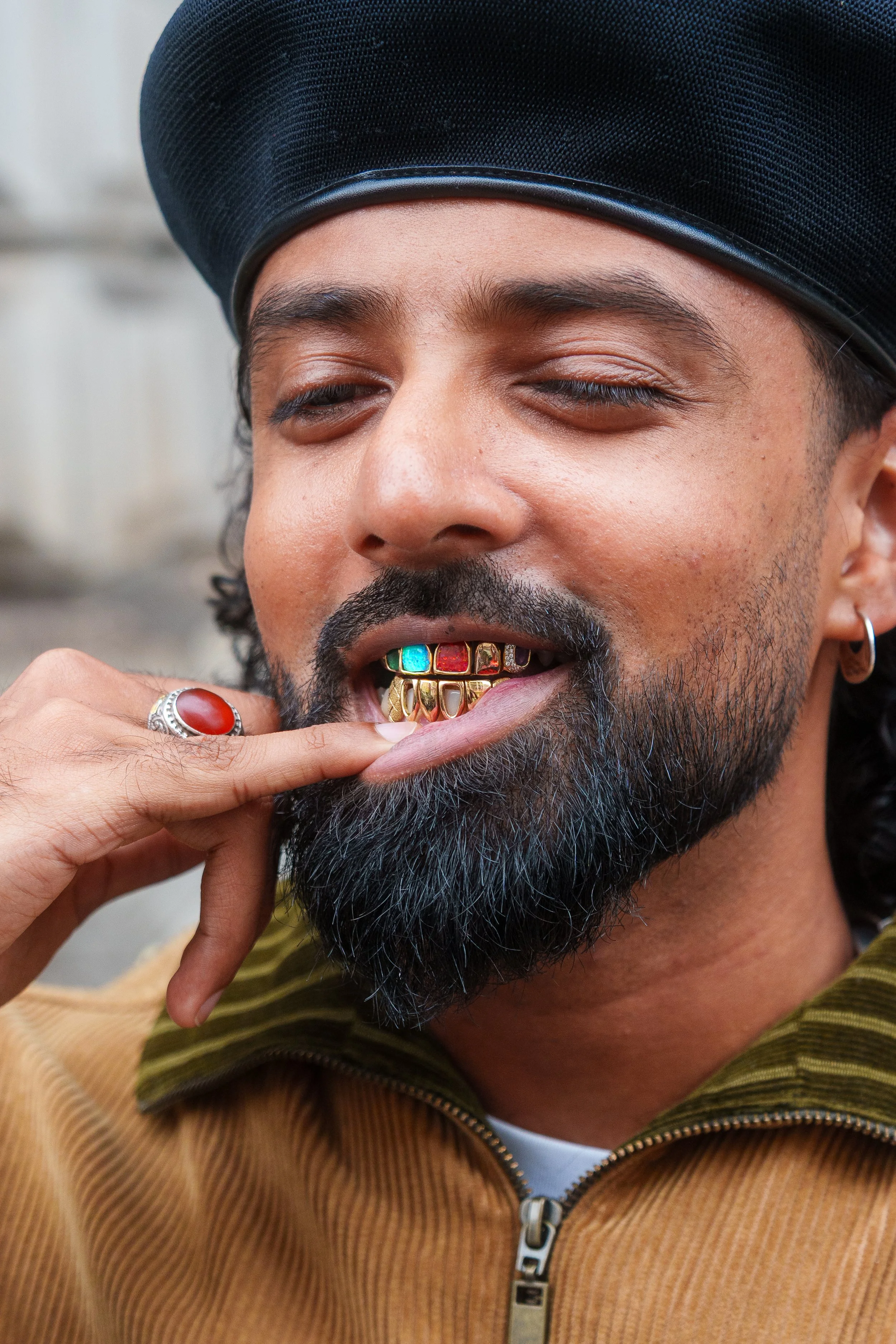 A man with dark skin, facial hair, and closed eyes is wearing a black hat and gold grillz with colorful inlays and is touching his tongue with his finger.