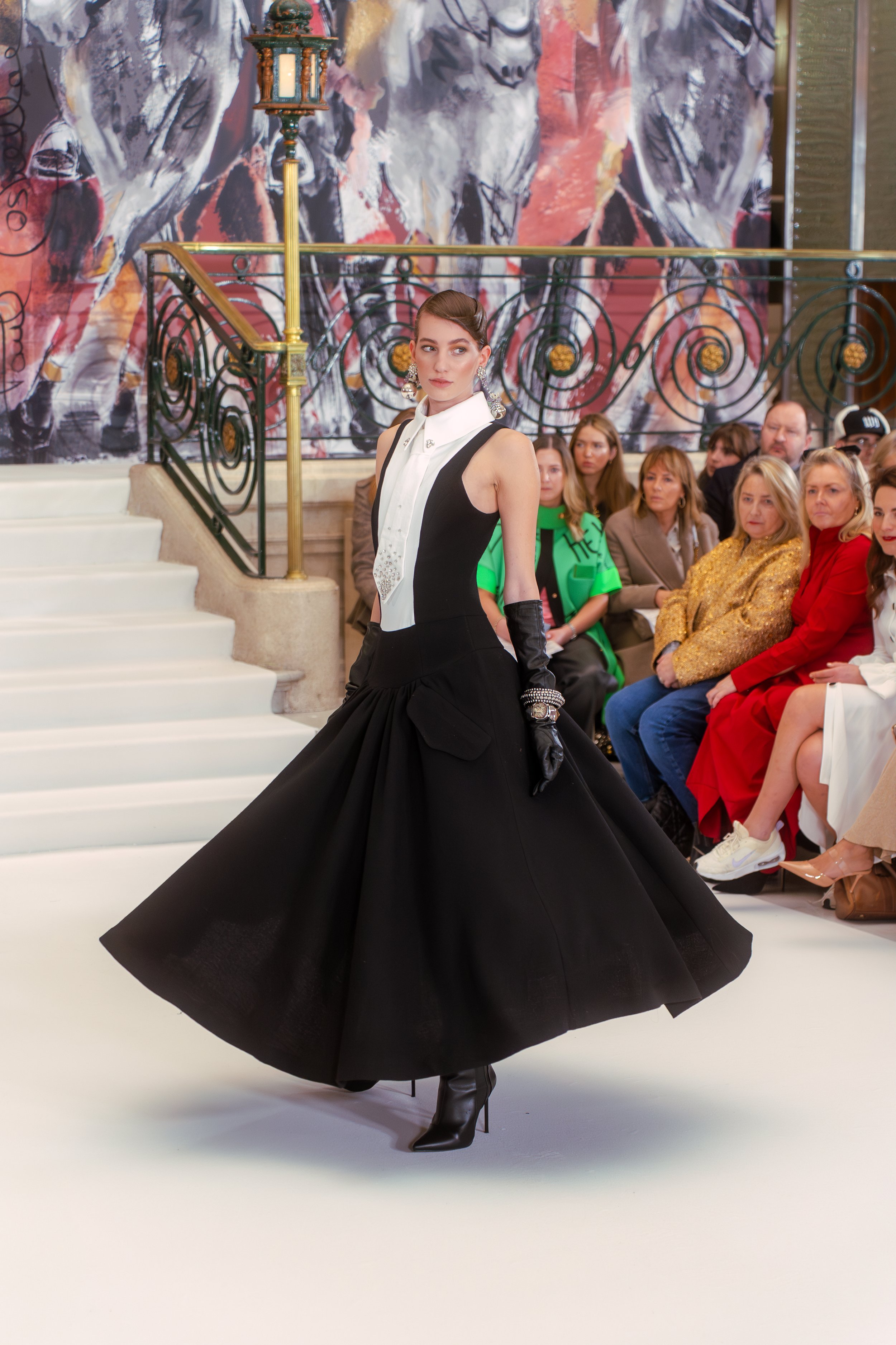 A model is walking on a runway during a fashion show, wearing a black and white high-fashion gown with a voluminous black skirt. She has dark lipstick, vintage-style hair, and long black gloves. Audience members are seated on the side, watching the model, with a large abstract painting in the background.