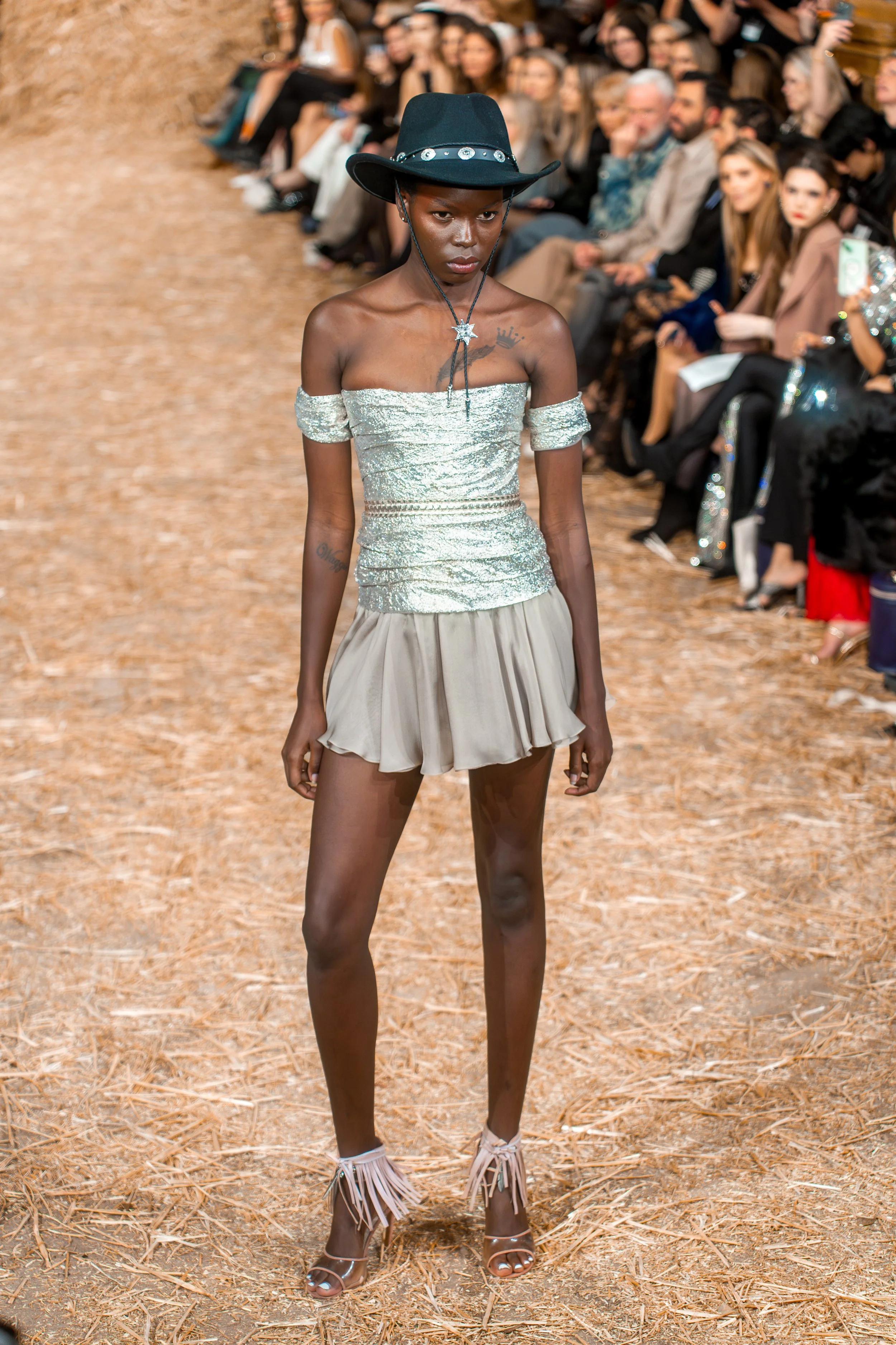 A female model walking on a runway during a fashion show, wearing a metallic off-shoulder top, a beige pleated skirt, a black hat with silver accents, and silver high heels with fringe details.