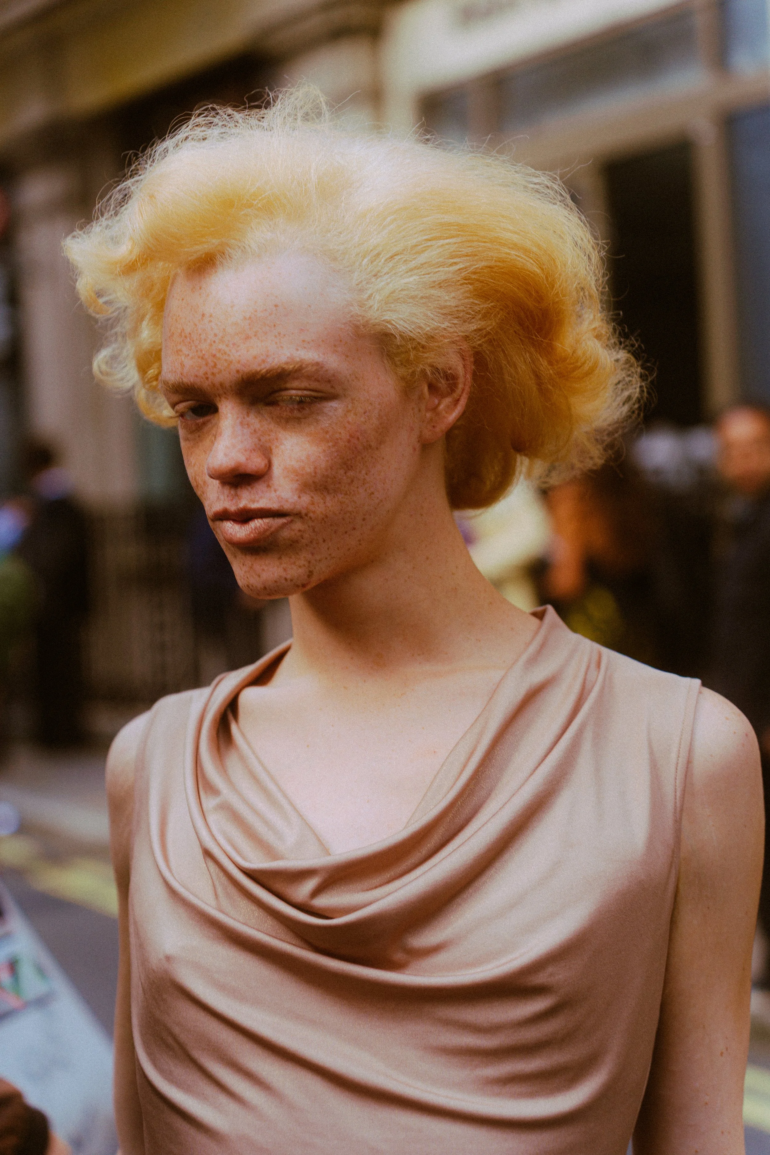 A woman with pale skin, freckles, and platinum blonde hair styled in loose waves, winking at the camera, wearing a beige satin sleeveless dress in an outdoor setting with blurred people in the background.