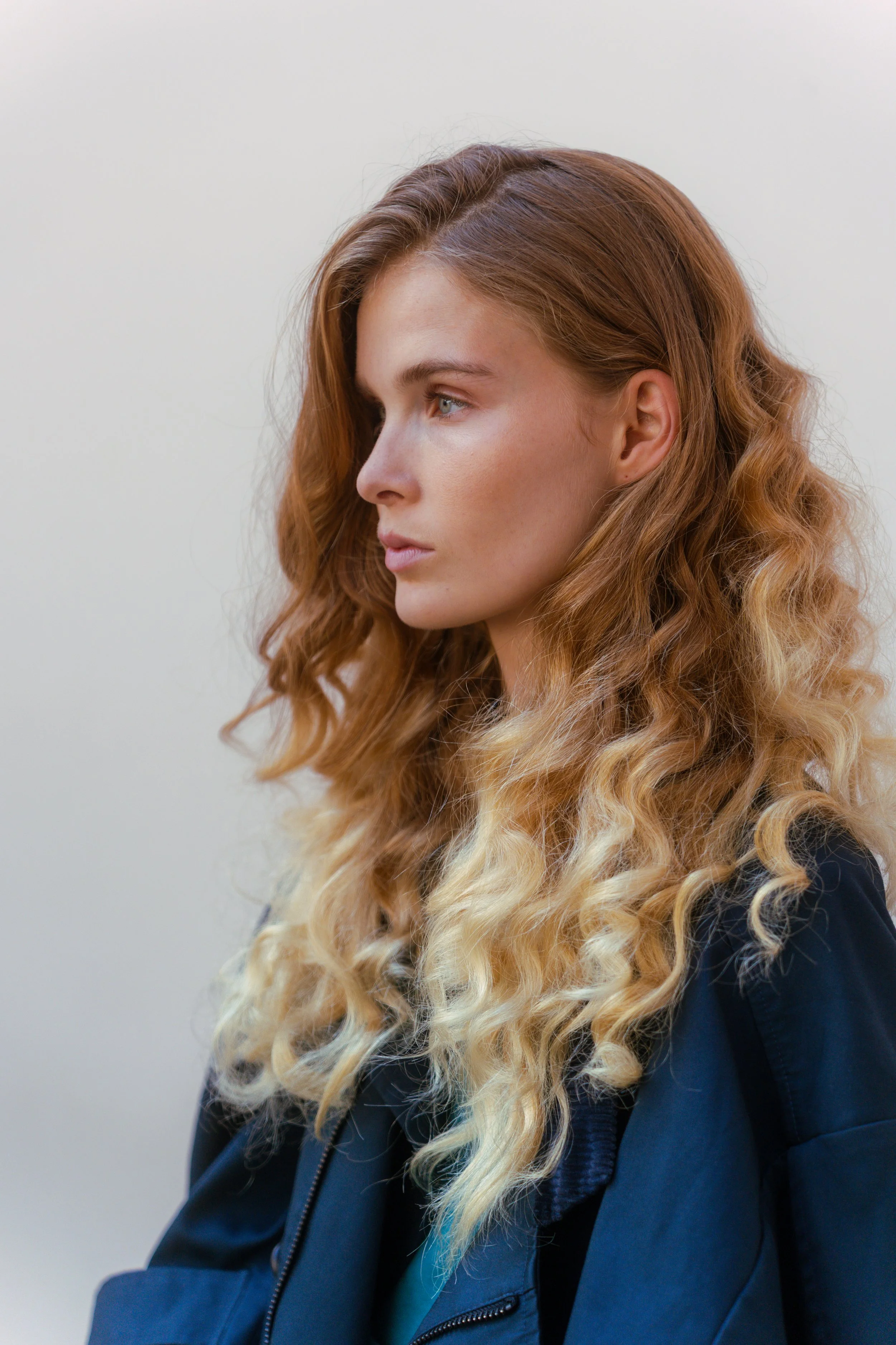 A woman with long, curly red hair and blue eyes looking to the left, wearing a black jacket, standing against a plain gray background.