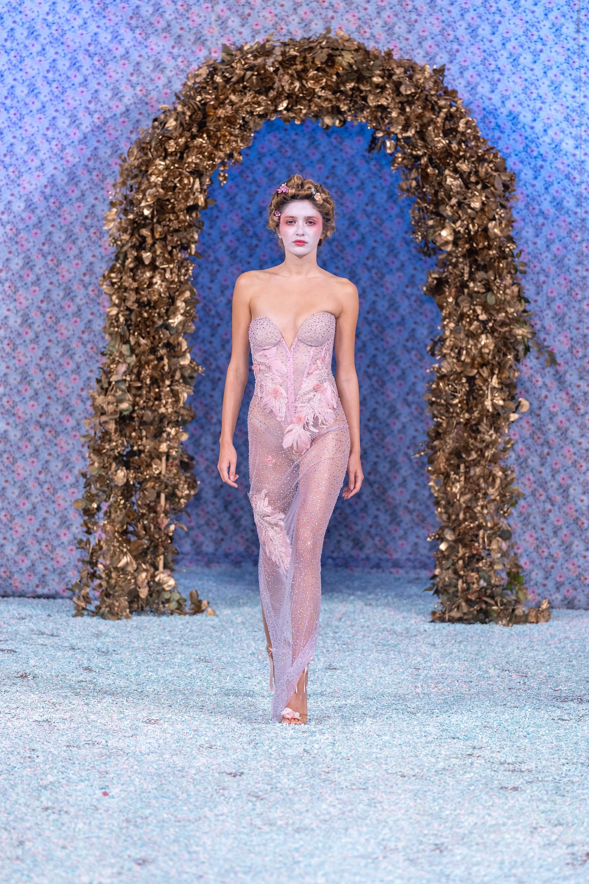 A fashion model walking on a runway under a floral arch with petals in pink and brown hues, wearing a strapless, sheer pink gown with floral appliqué and intricate beadwork, set against a textured blue background.