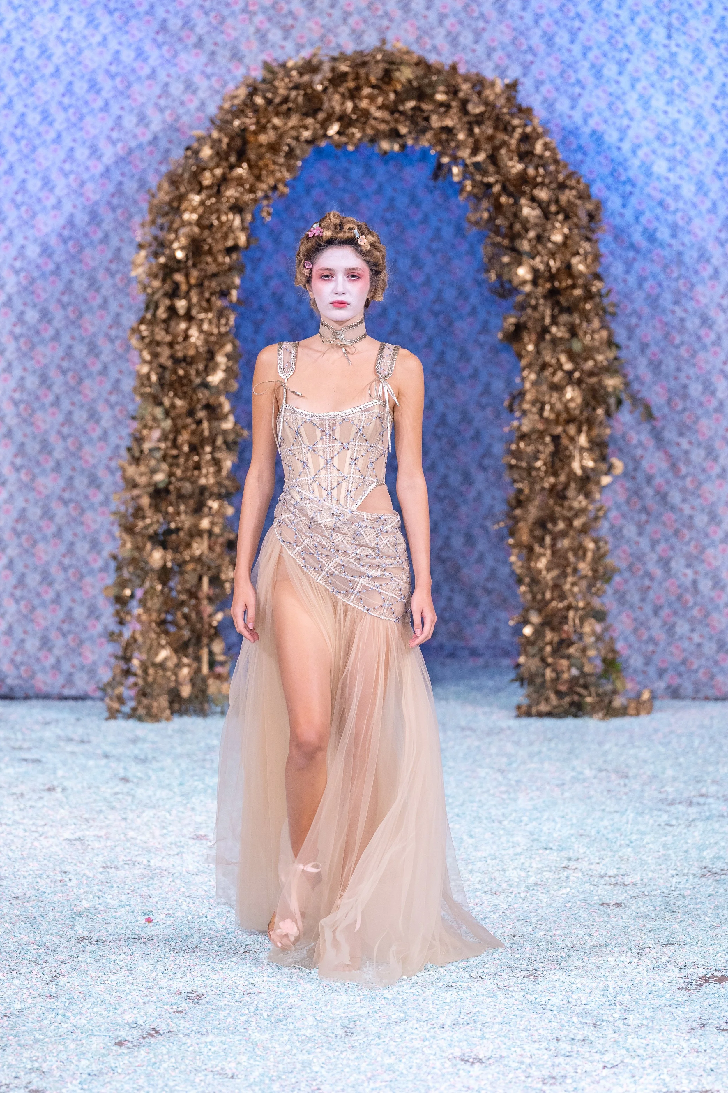 Model walking on runway in beige gown with sheer skirt and intricate beaded bodice, backdrop features a floral patterned wall and arch decorated with gold-colored leaves.