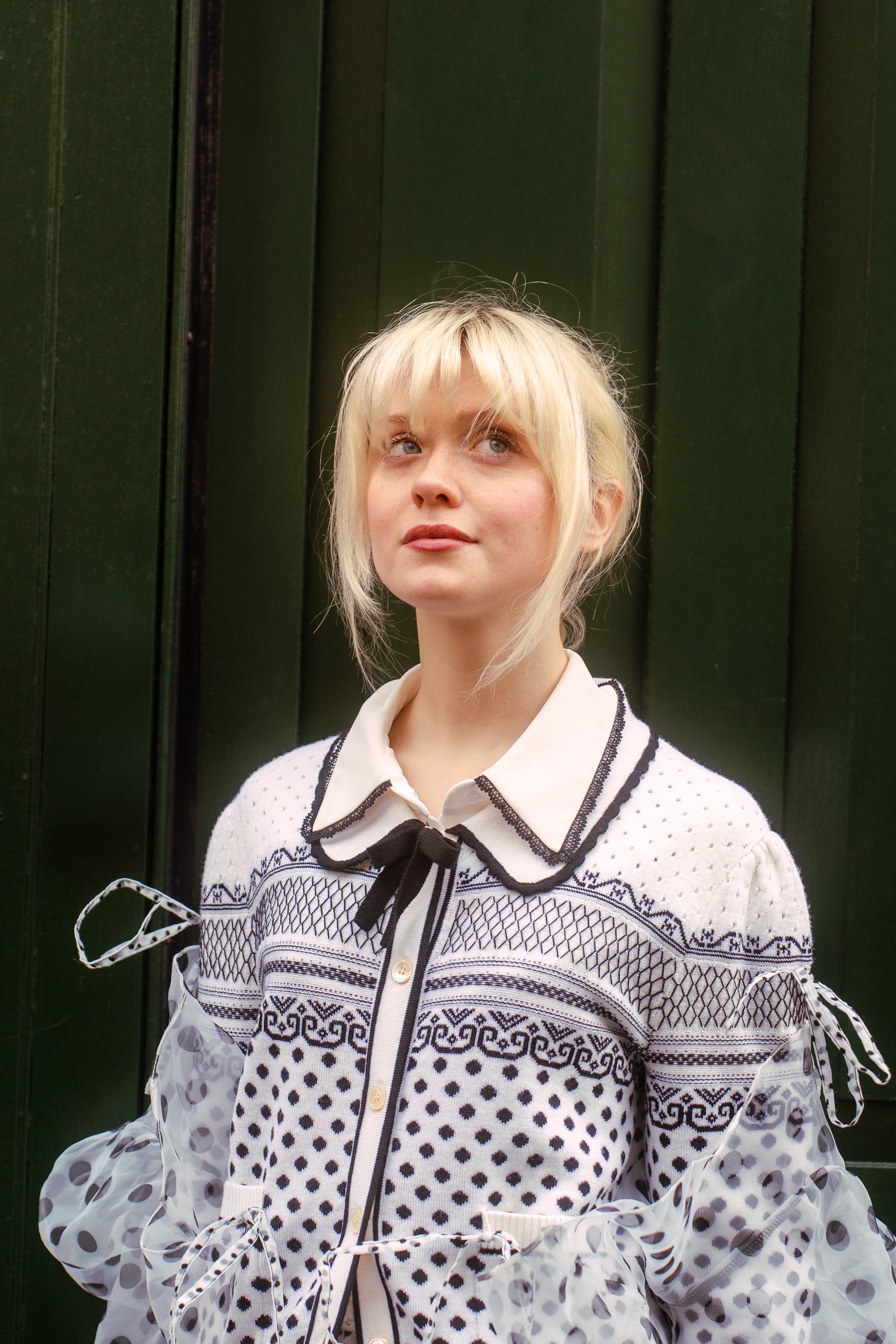 A young woman with blonde hair, wearing a white shirt with a black bow tie, and a cardigan with intricate black and white patterns. She stands against a dark green wooden wall.