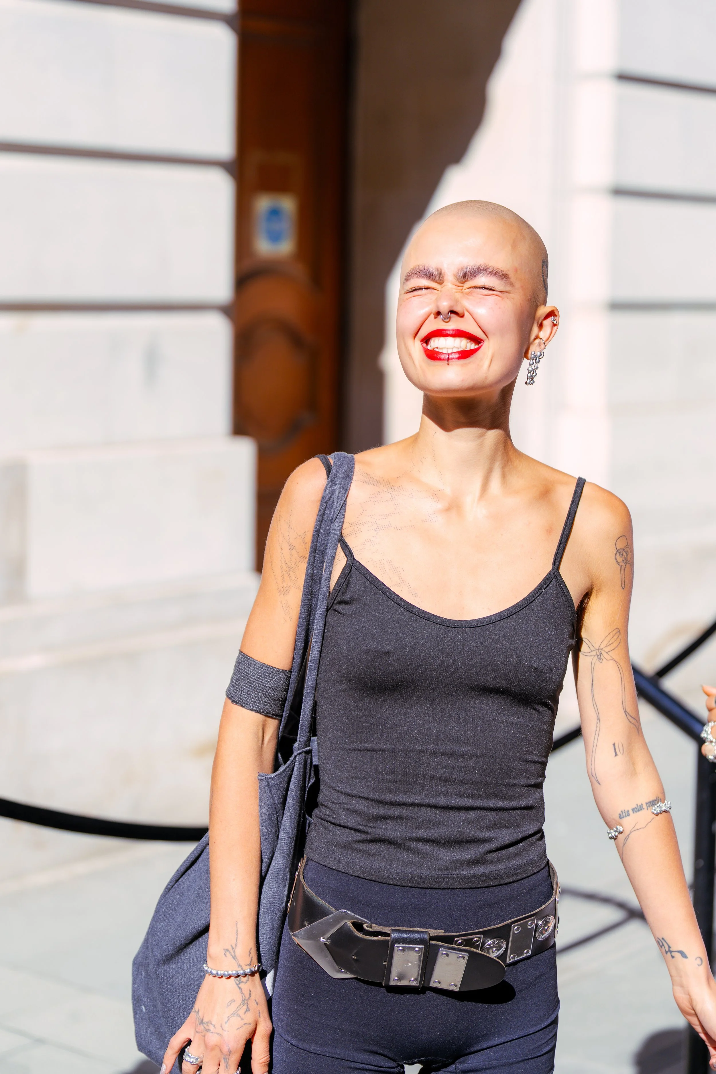 A smiling woman with a shaved head, wearing a black tank top, jewelry, and carrying a gray bag, standing outdoors in bright sunlight.