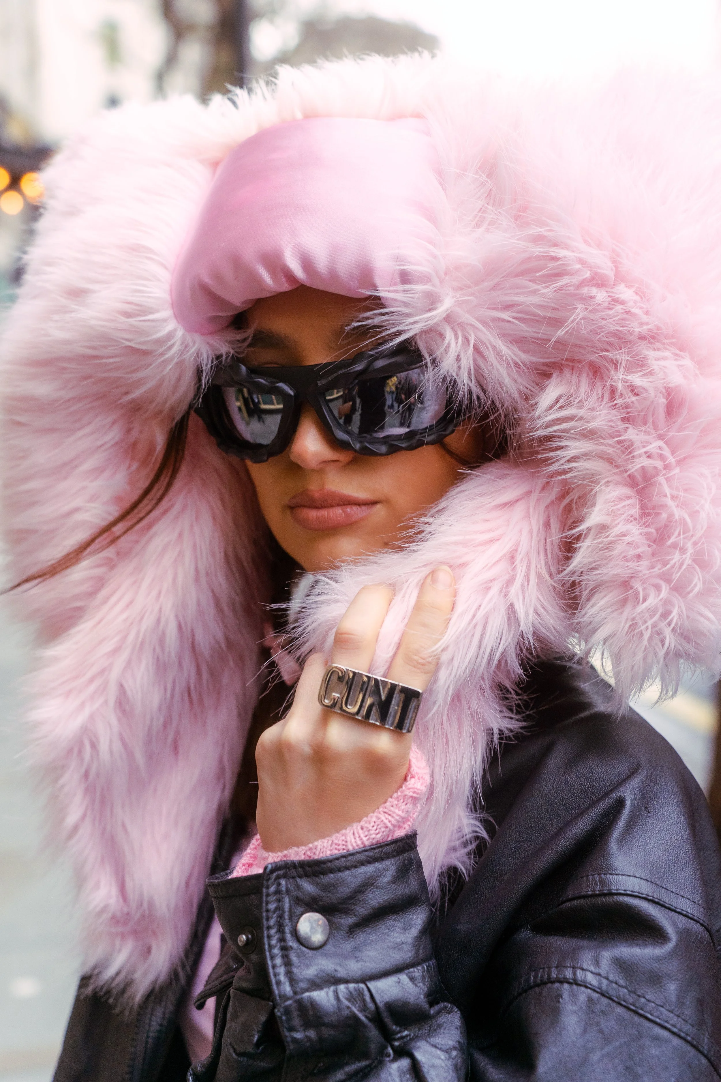 Woman wearing large pink furry hooded coat, black leather jacket, and black sunglasses with a ring that says 'GUNTI' on her finger.