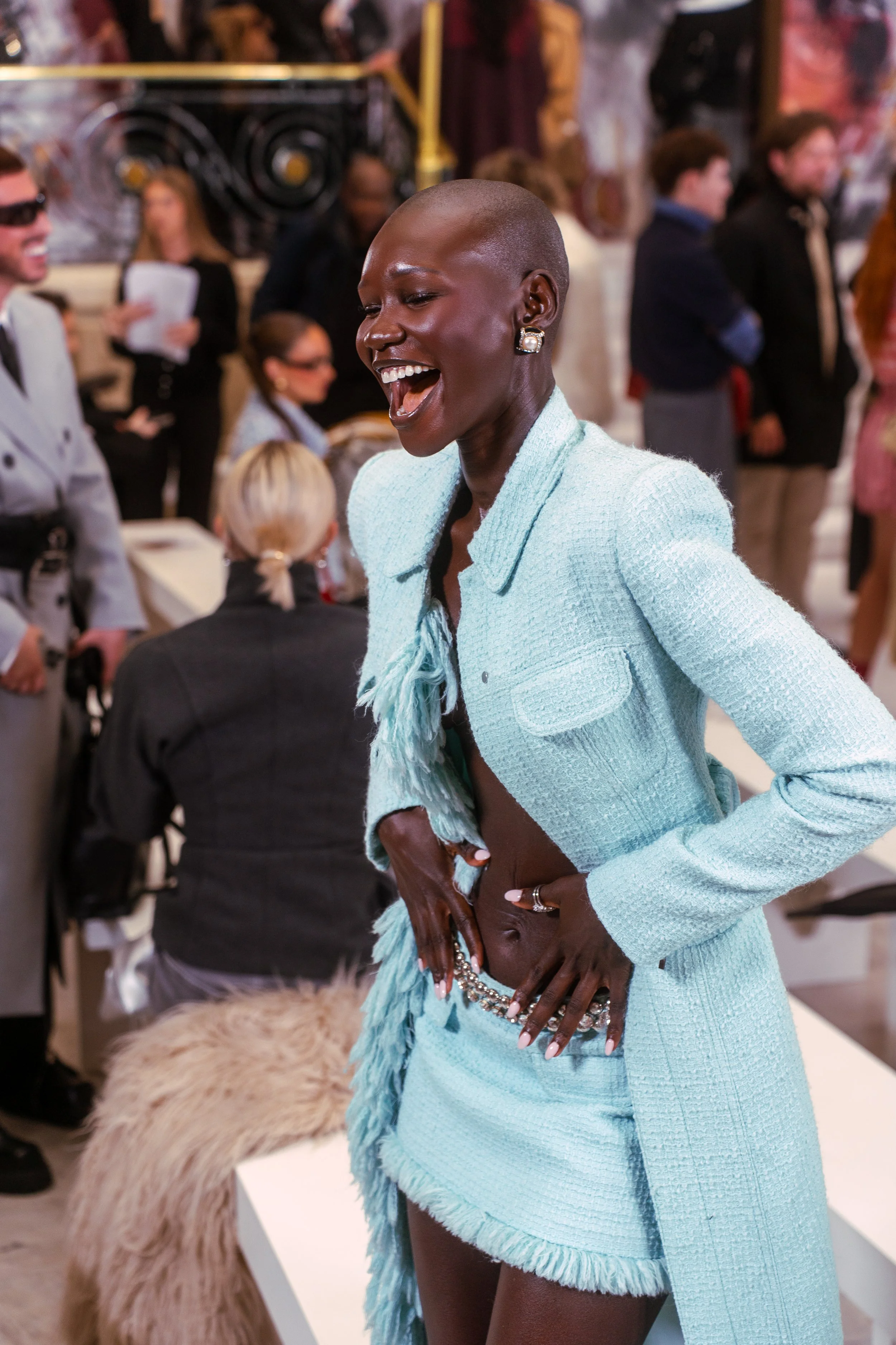 A woman in a light blue textured jacket and matching skirt is smiling and laughing at a social event, with other people in the background.