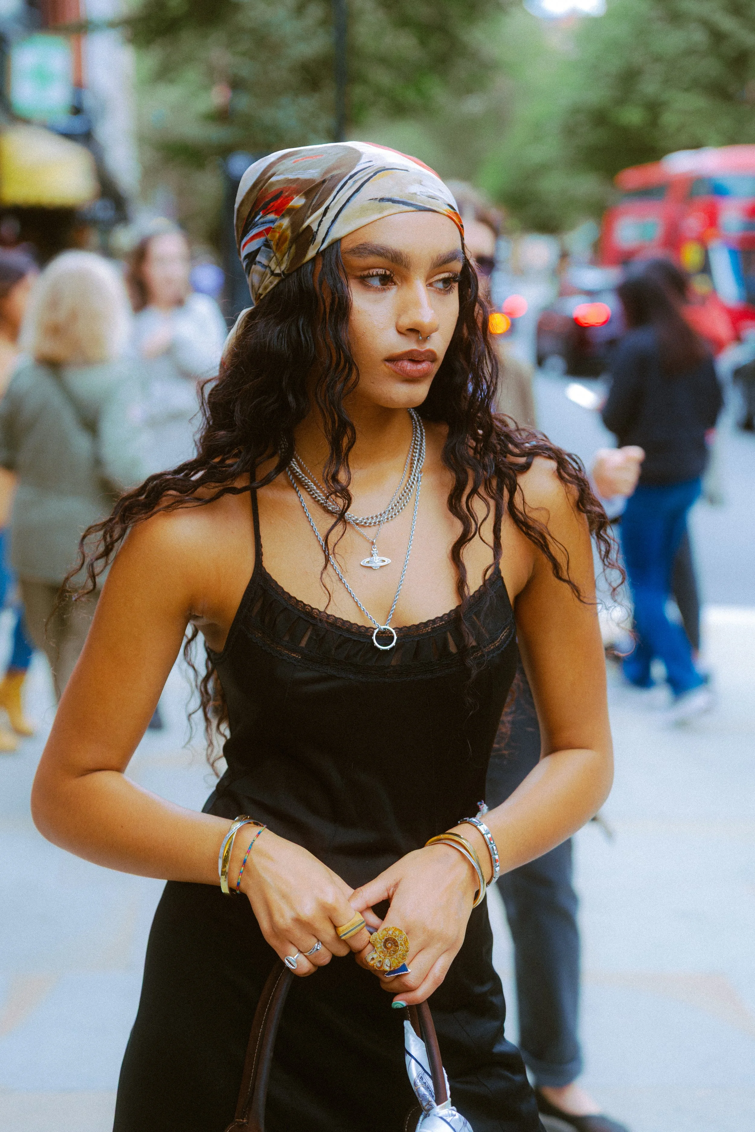A young woman with long dark curly hair wearing a patterned headscarf, multiple layered necklaces, rings, and bracelets, dressed in a black camisole, standing on a busy city street with people and vehicles in the background.