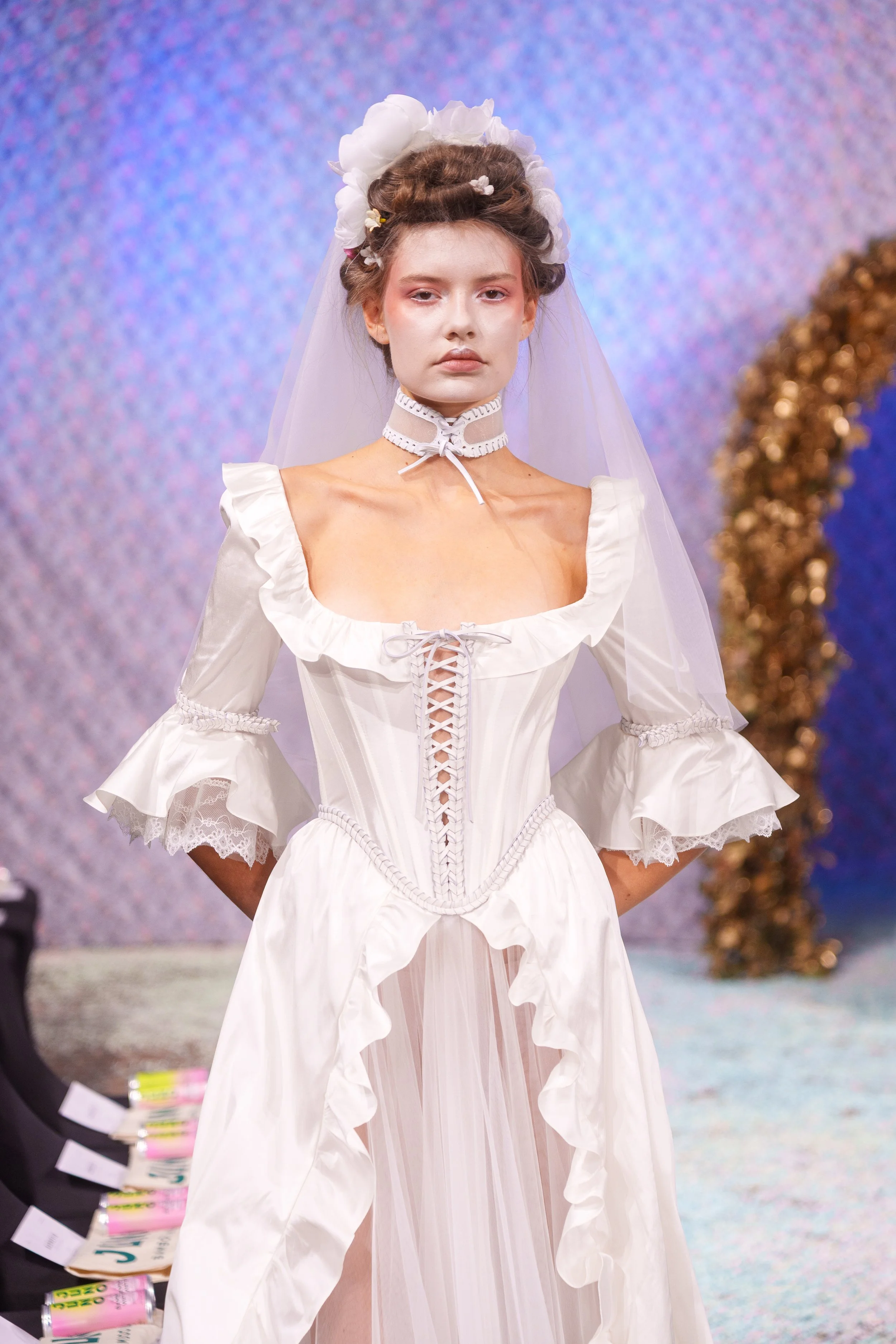 A woman walking on a runway in a white wedding dress with puffed sleeves, lace-up front, ruffled details, and a sheer overlay. She is wearing a floral headpiece, a veil, and a lace choker, with her hands behind her back and a serious expression.