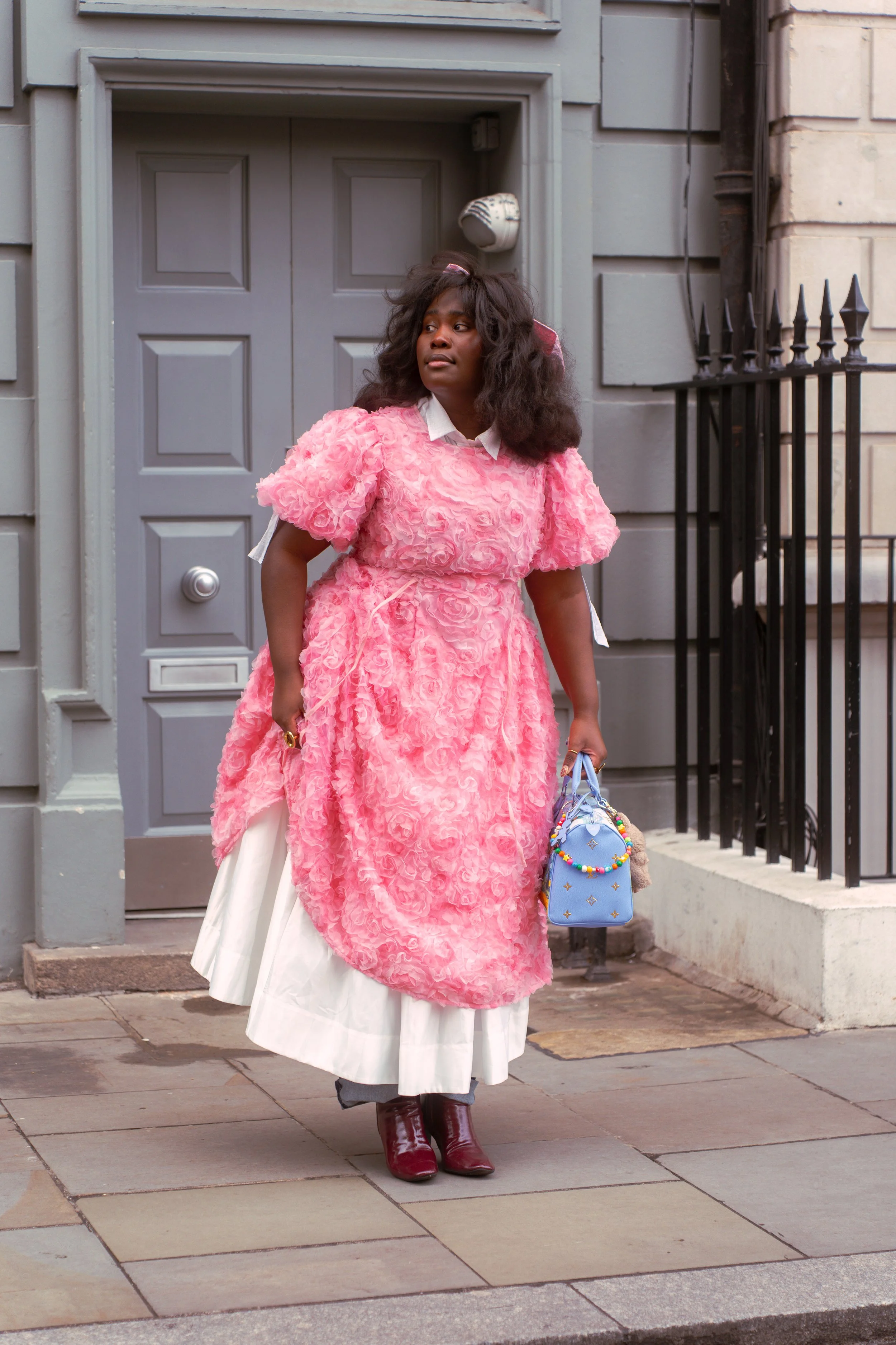 A woman dressed in a pink textured dress with white layers underneath, holding a small blue handbag, standing on a sidewalk in front of a gray building with a door and black iron railing.