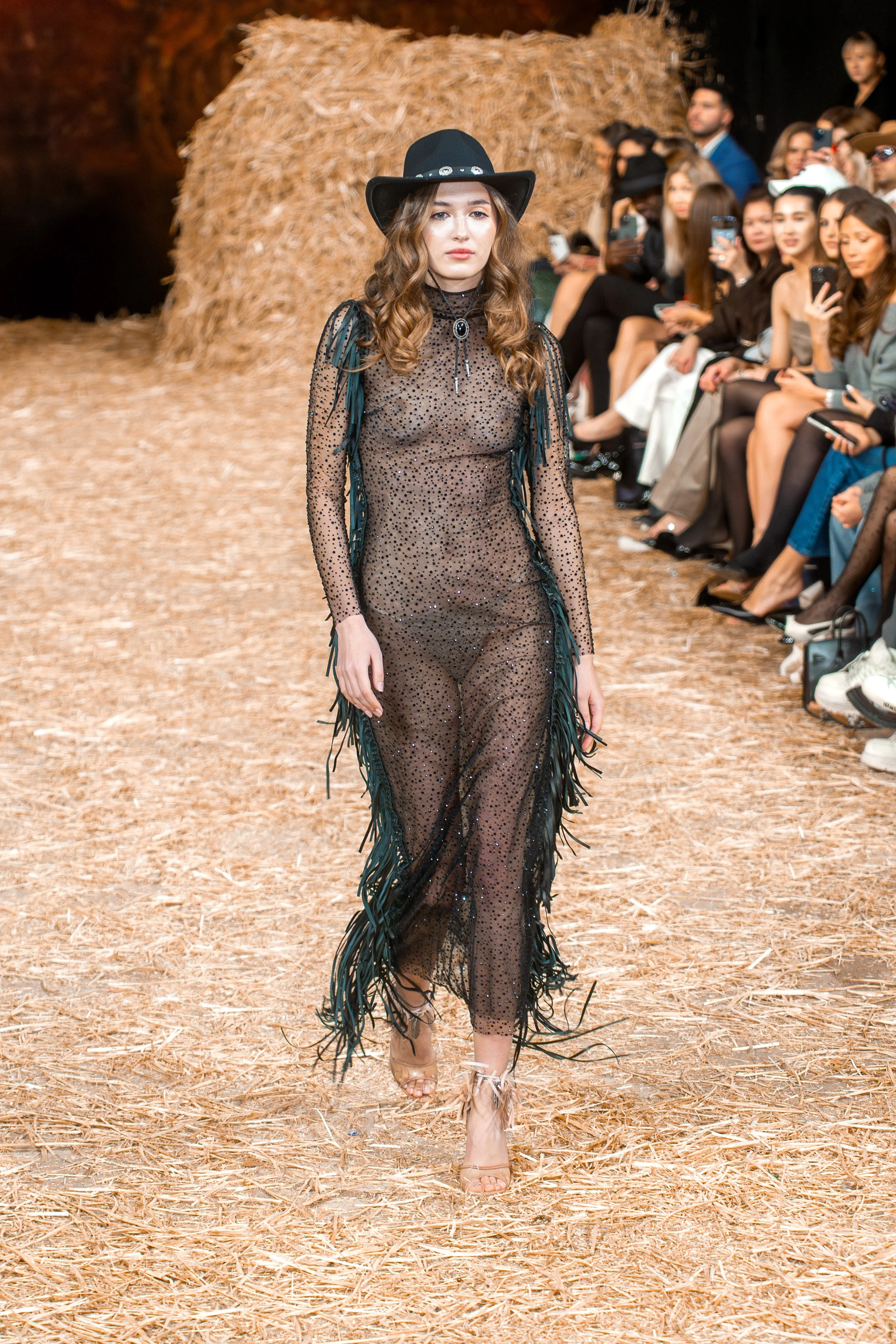 A model walking down a runway covered with straw, wearing a sheer black dress with small sequins and fringe details, paired with a black cowboy hat, with an audience watching on the sides.