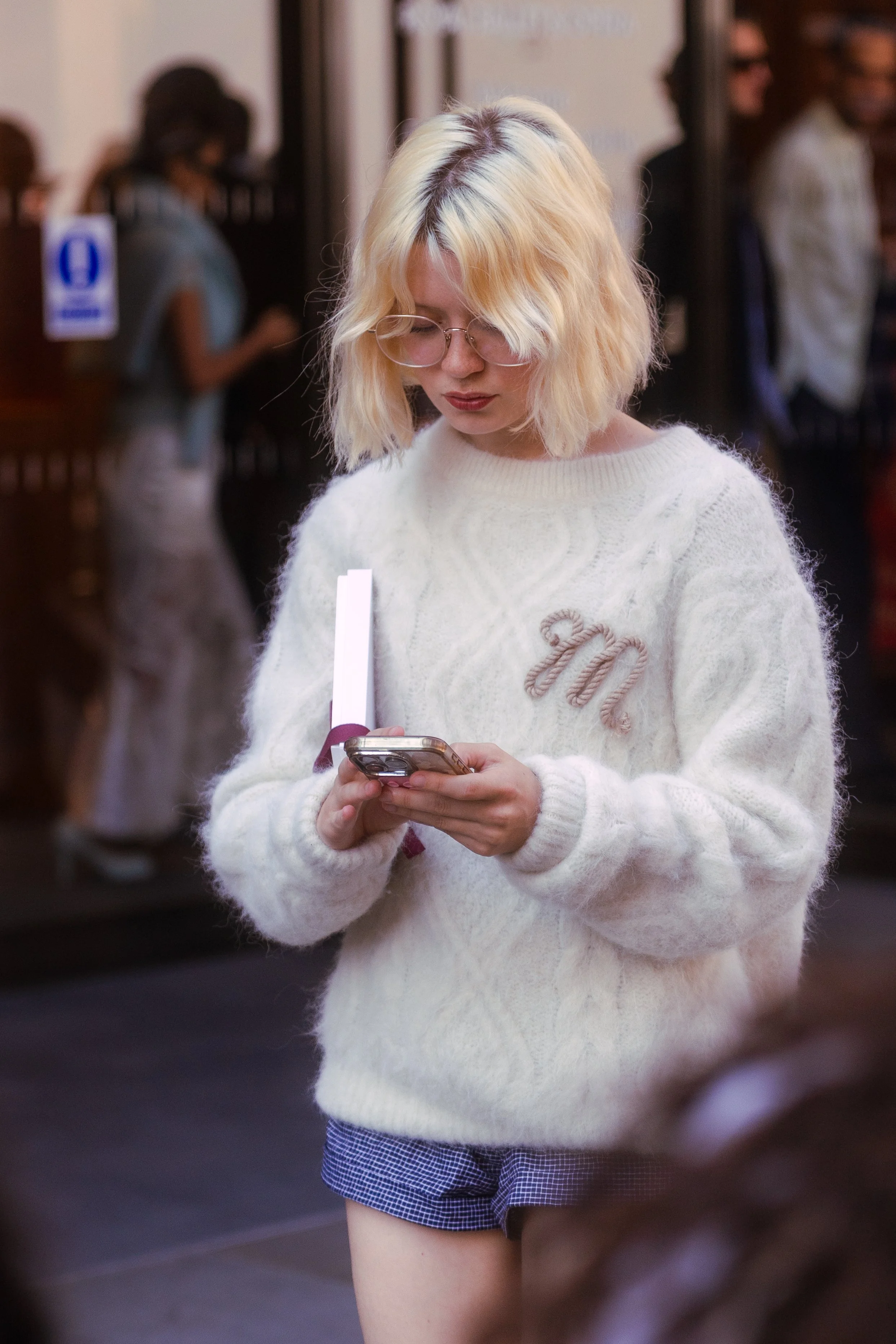 Young woman with blond hair, glasses, and a white sweater looking at her phone in a busy indoor setting.