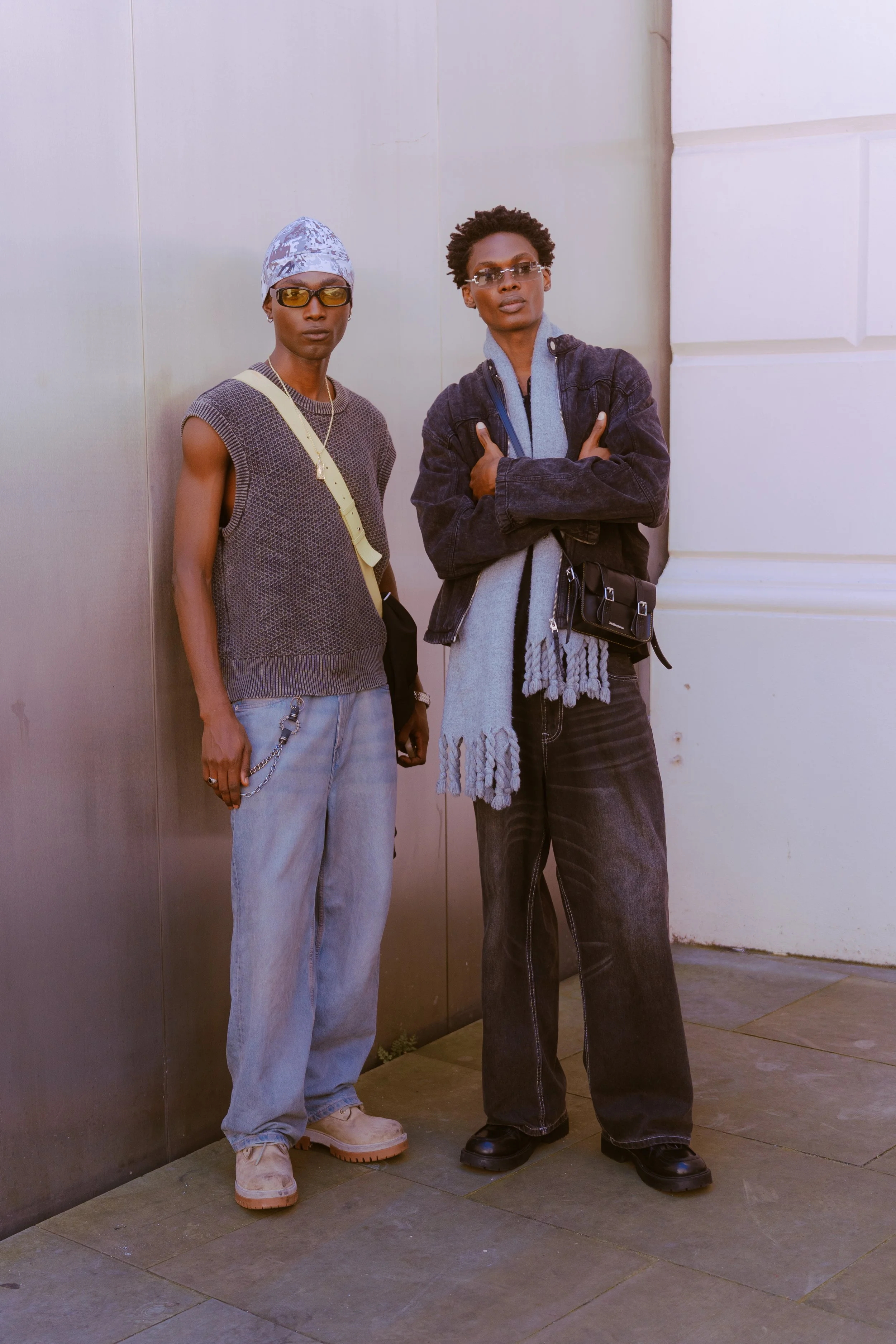 Two individuals with dark skin wearing sunglasses, standing against a wall, dressed in casual, vintage-inspired clothing.