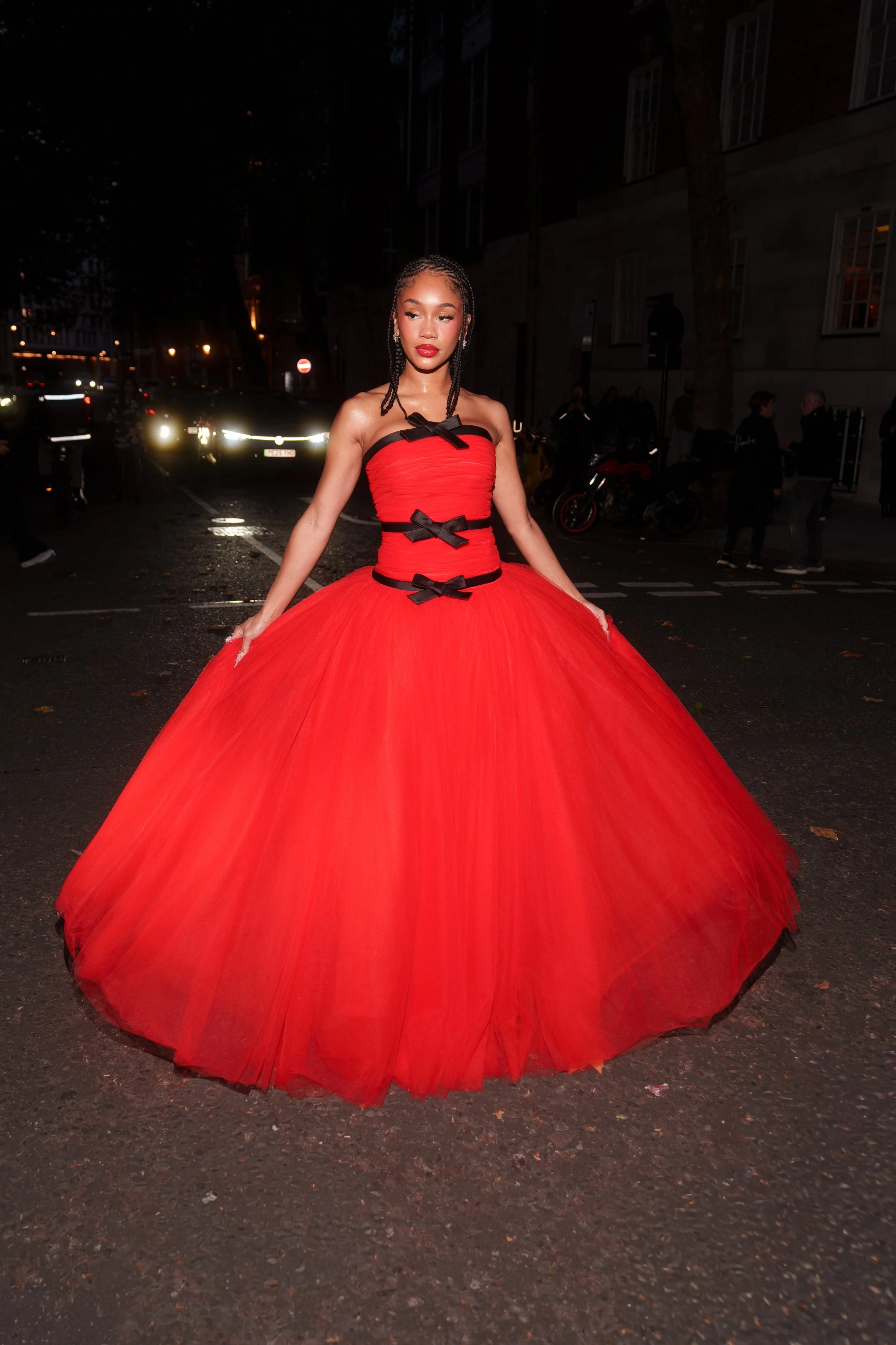 A woman in a red strapless ball gown with black bows, standing on a city street at night.