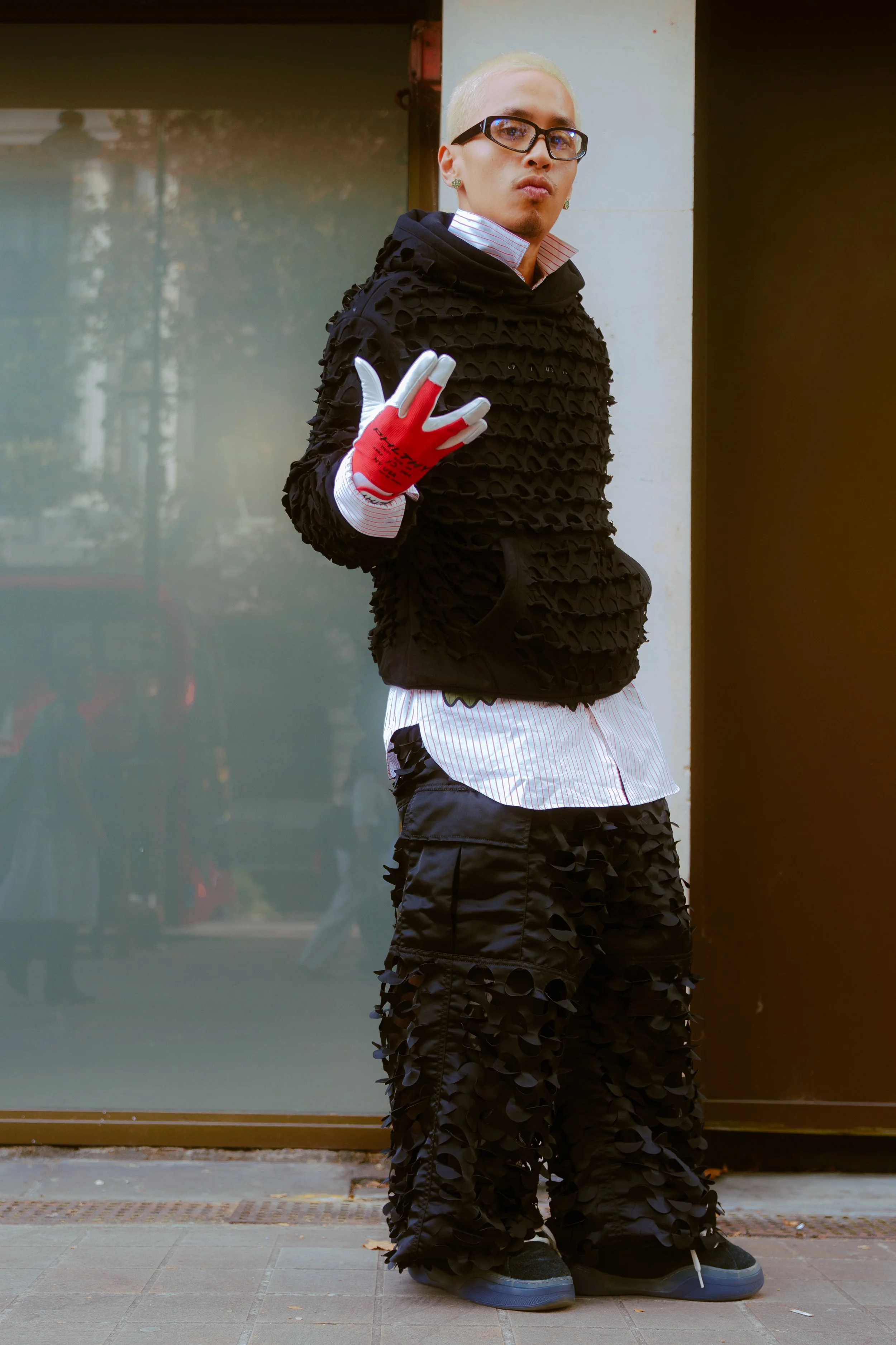 Person with platinum blonde hair, wearing glasses, a black textured hoodie, white striped shirt, black textured pants, and black shoes, standing outdoors with one hand raised giving a peace sign.