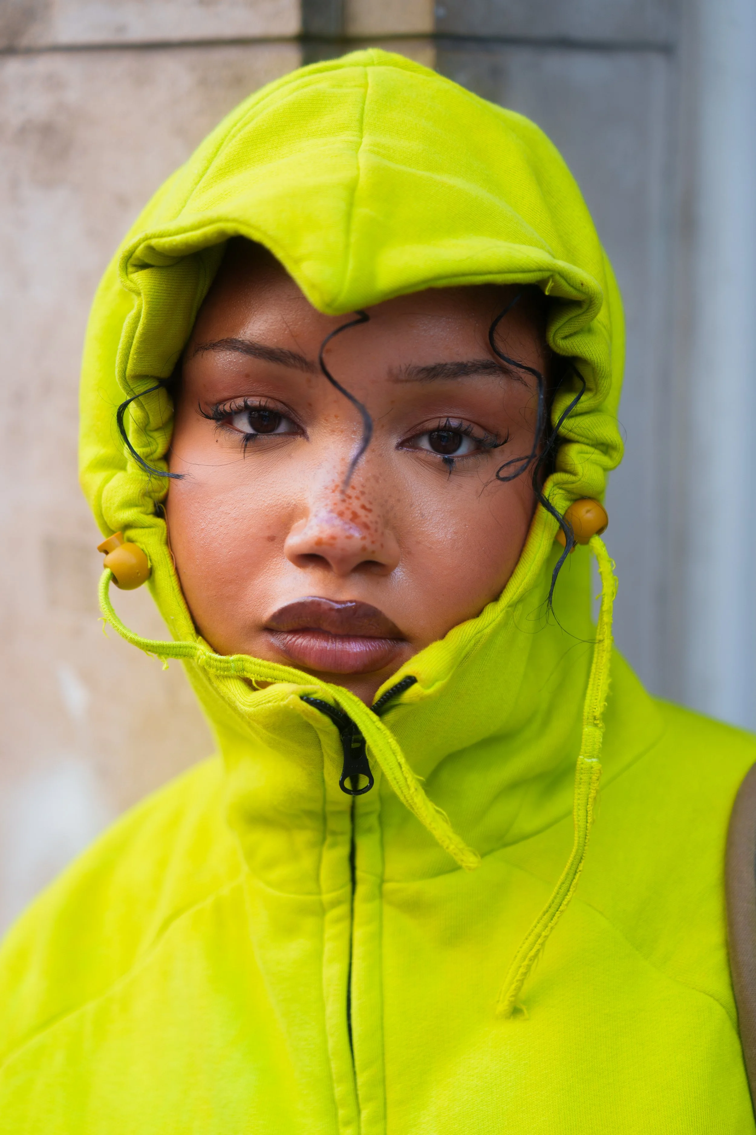 A young woman with curly black hair and freckles, wearing a bright yellow hoodie with the hood over her head, standing outdoors near a concrete wall.