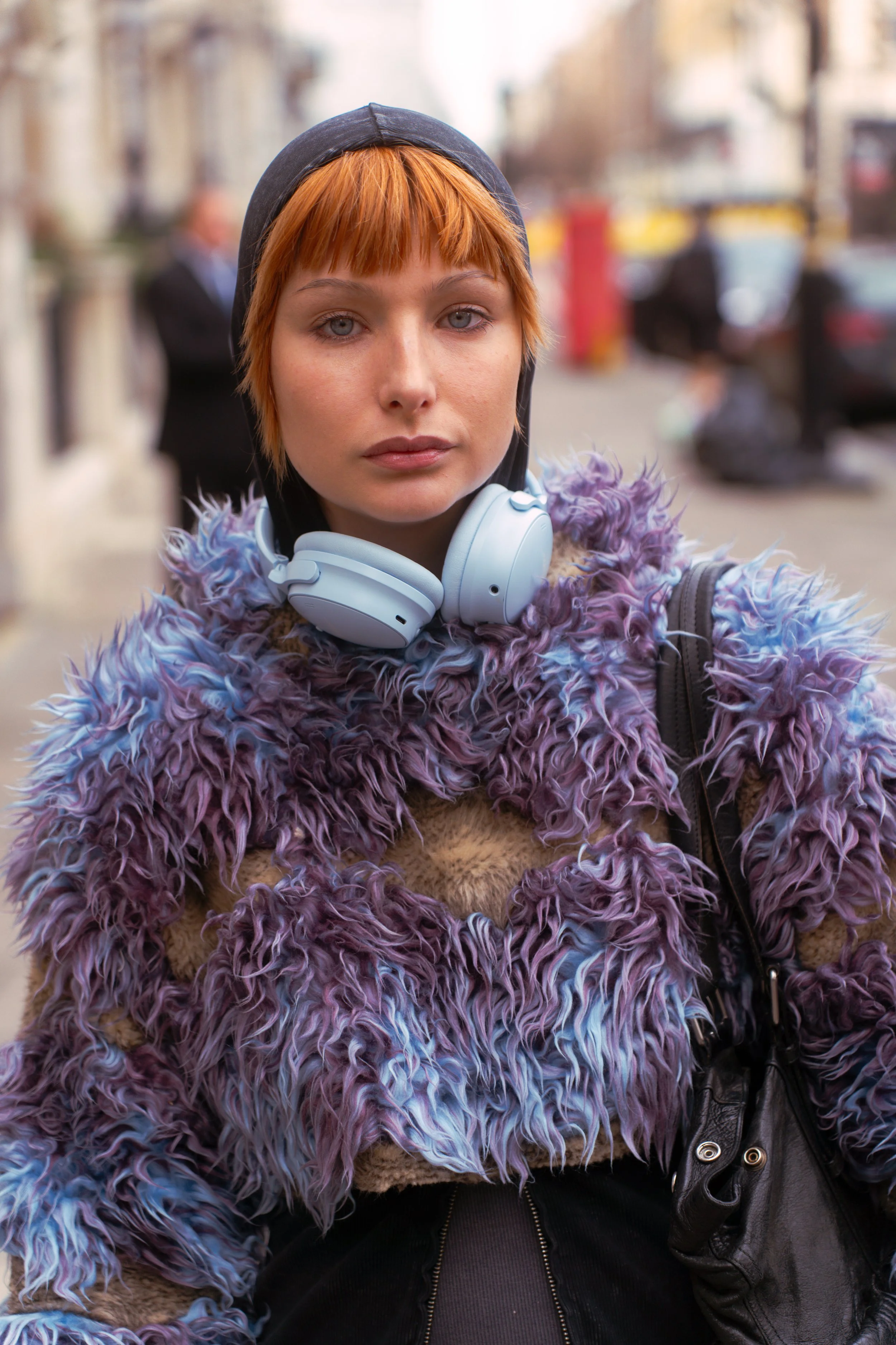 Young woman with short red hair wearing a furry purple and blue coat, black hoodie, and white headphones around her neck, standing outdoors on a city street.