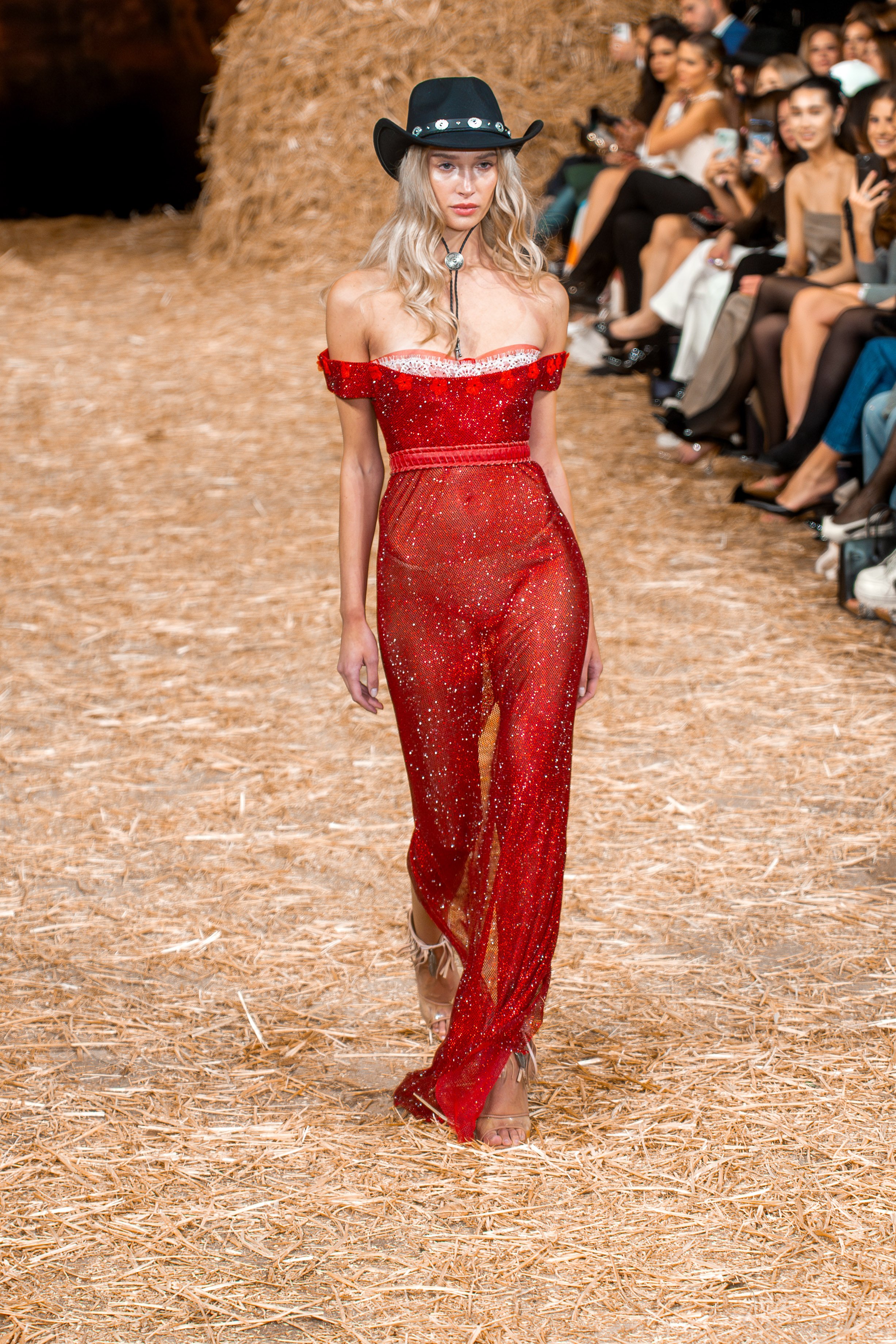 A female model walking down the runway at a fashion show, wearing a red sparkle off-shoulder jumpsuit and a black cowboy hat, with audience members seated on either side of the runway.
