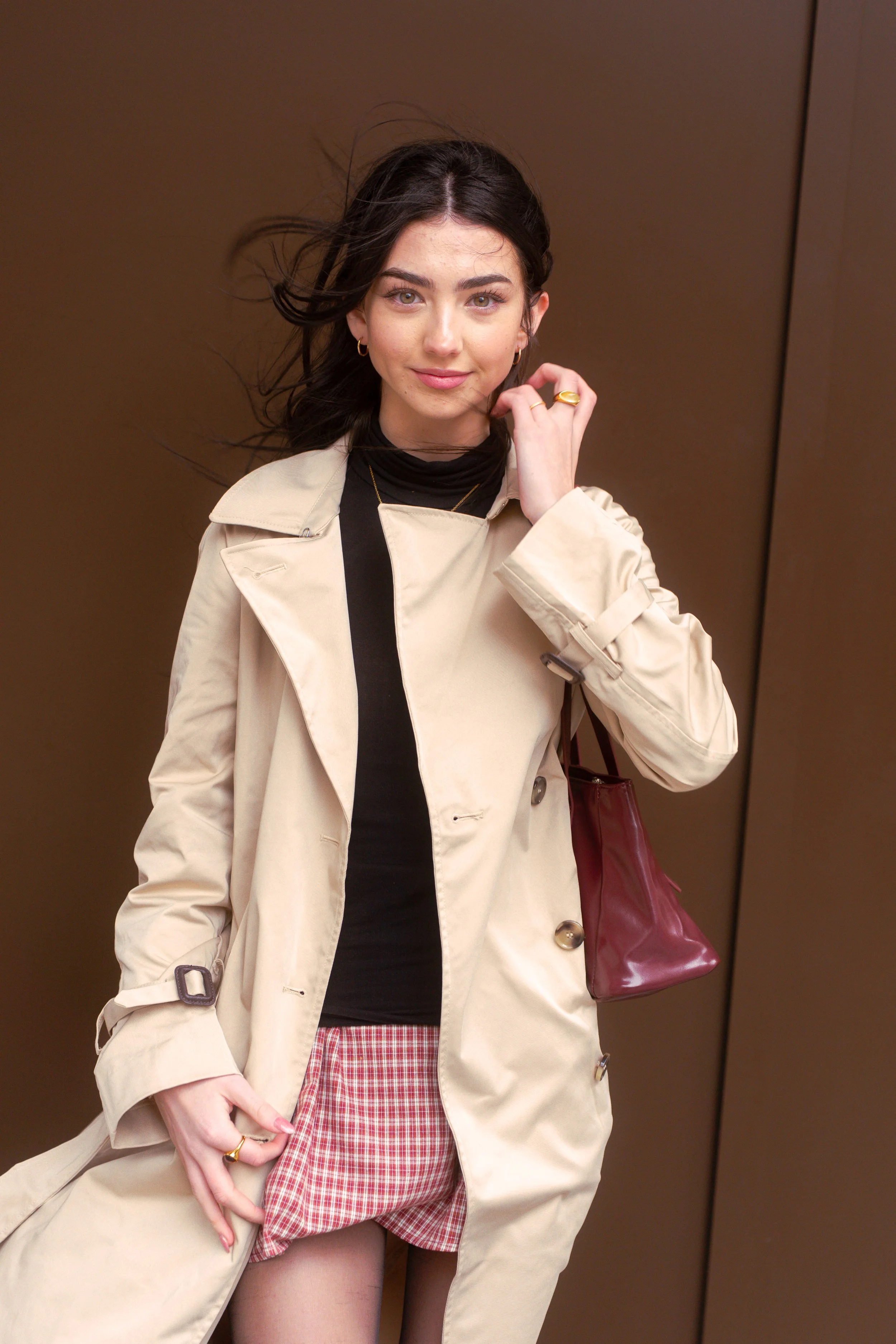Woman in beige trench coat, black turtleneck, red plaid shorts, holding a maroon bag, standing in an elevator