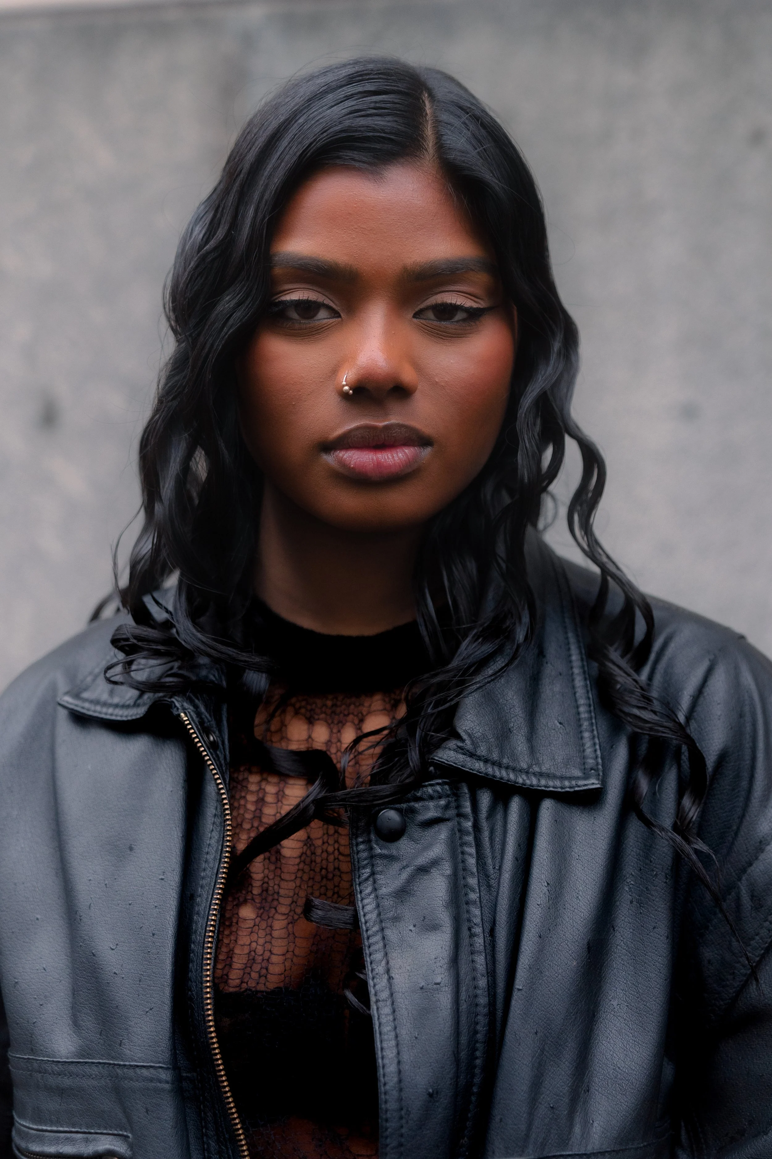A young woman with dark, wavy hair and dark skin wearing a black leather jacket and black lace top, standing against a plain gray background.