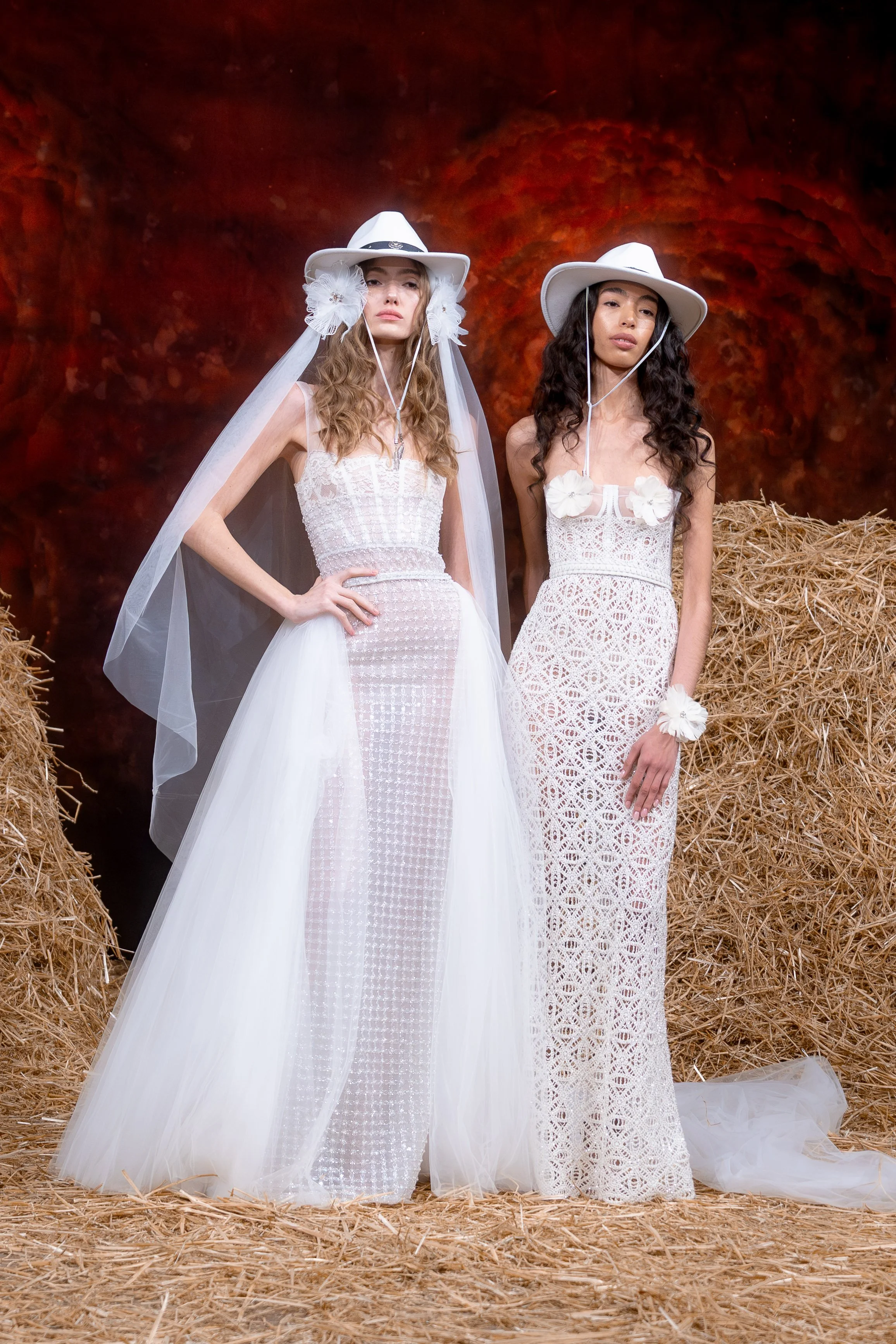 Two models in white wedding dresses with wide-brimmed hats posing in a rustic hay setting with a red rock backdrop.