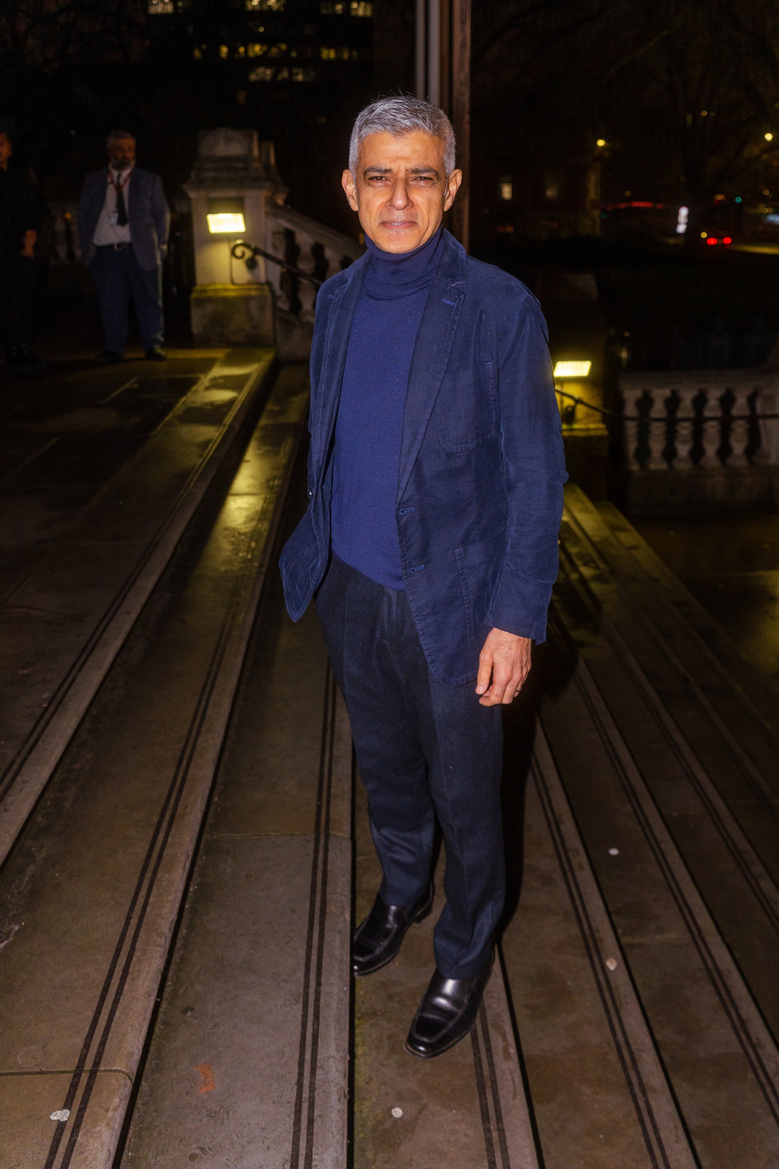 A man with gray hair wearing a dark blue turtleneck and blazer, dark pants, and shiny black shoes standing on a tram track outdoors at night.