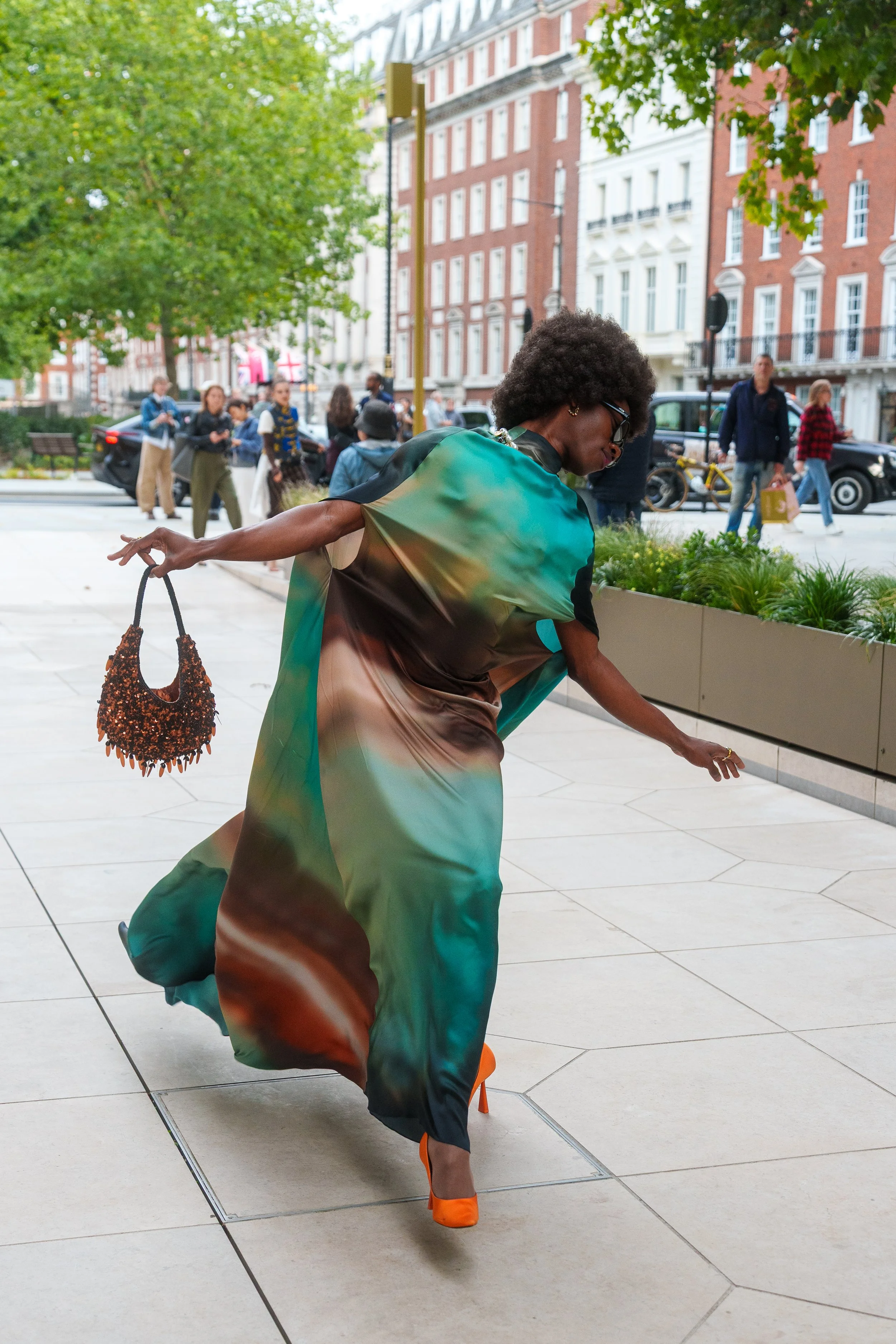 A woman in a colorful, flowy dress and bright orange heels is dancing or posing on a city sidewalk. She is holding a beaded handbag and glasses, with trees and a row of buildings and pedestrians in the background.