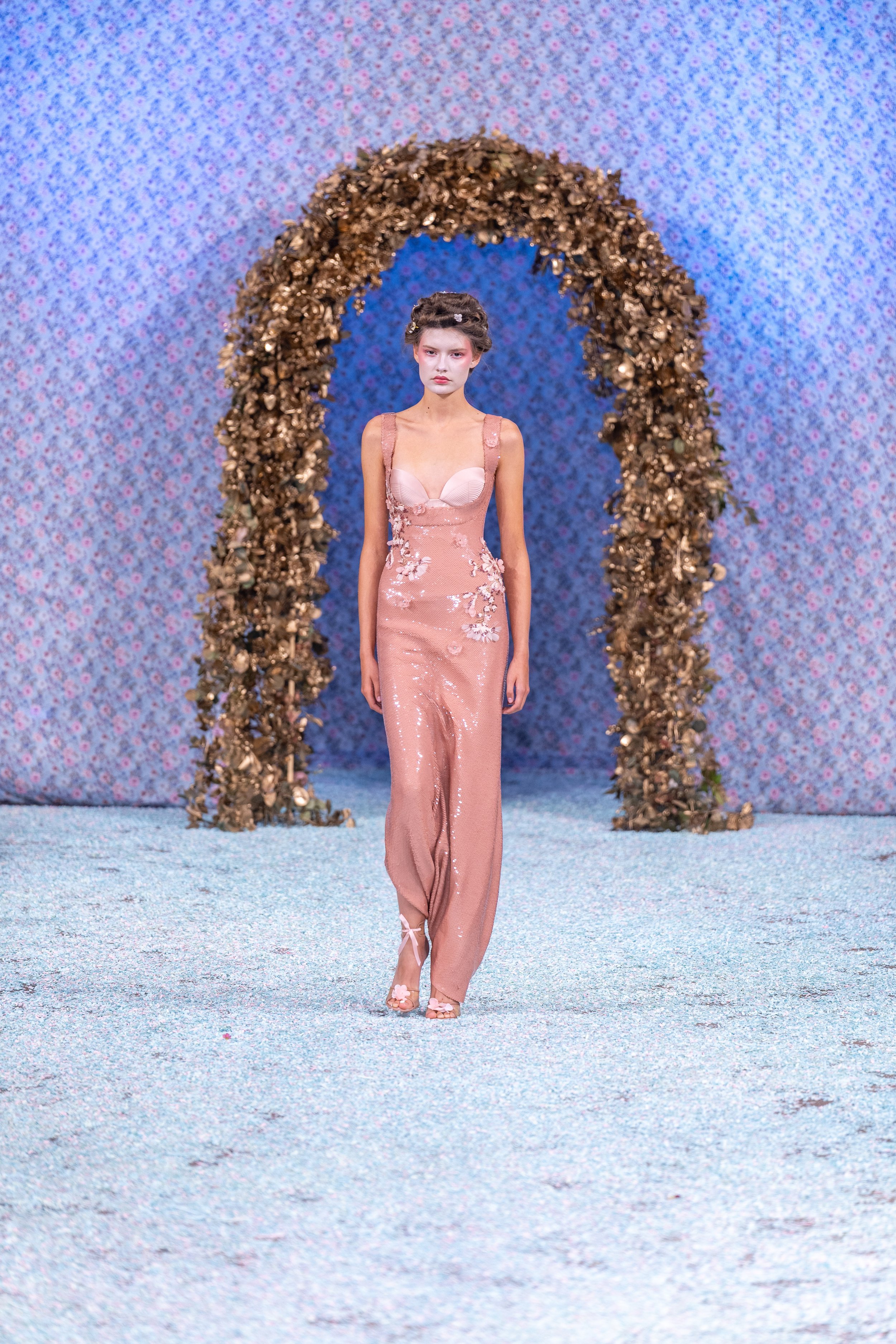 A model walks on a runway with a floral arch behind her and a pastel-colored background.