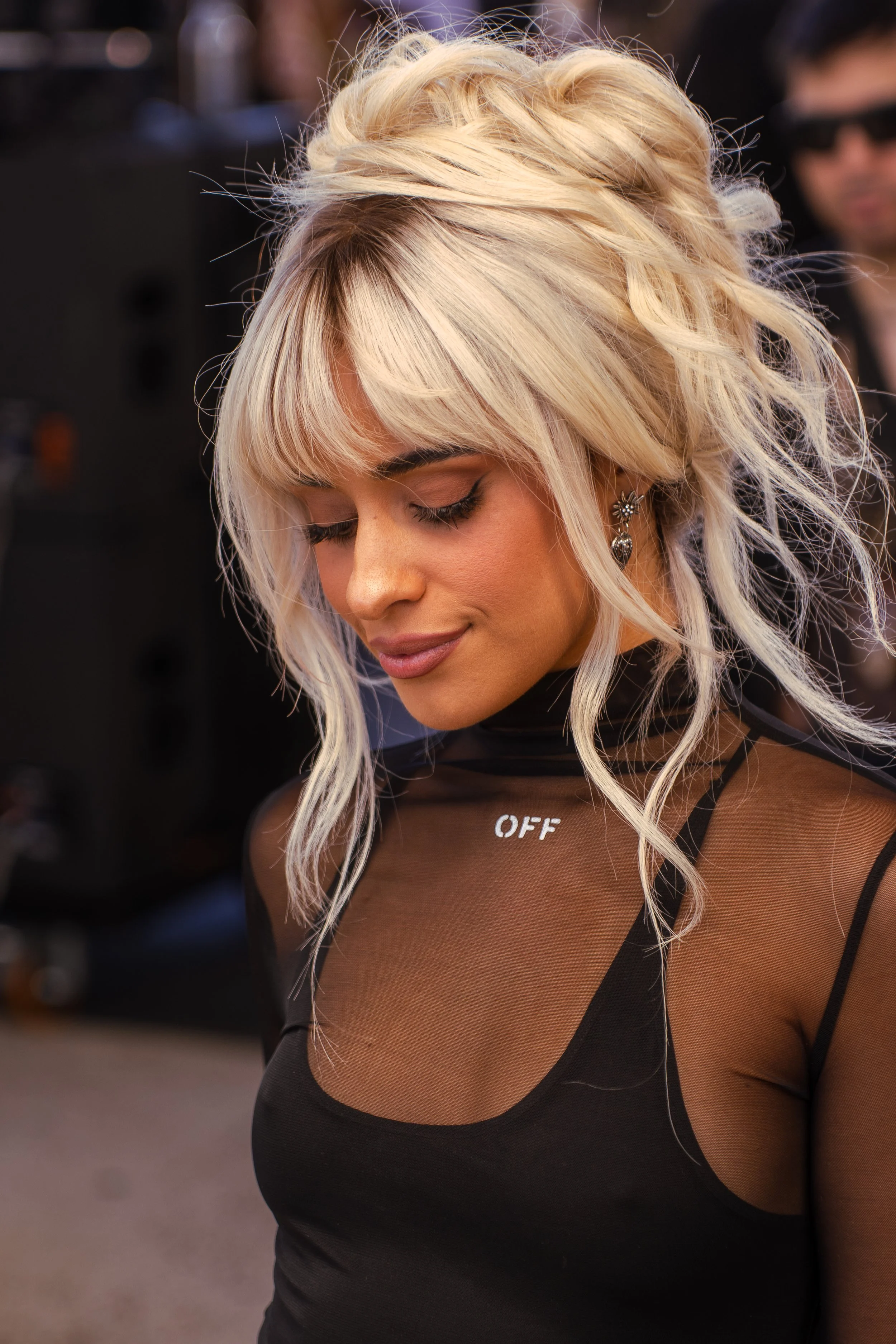 A woman with platinum blonde, wavy hair styled with loose, tousled waves and bangs, wearing earrings and a sheer black top with the word "OFF" on the collar, looking down with a slight smile.