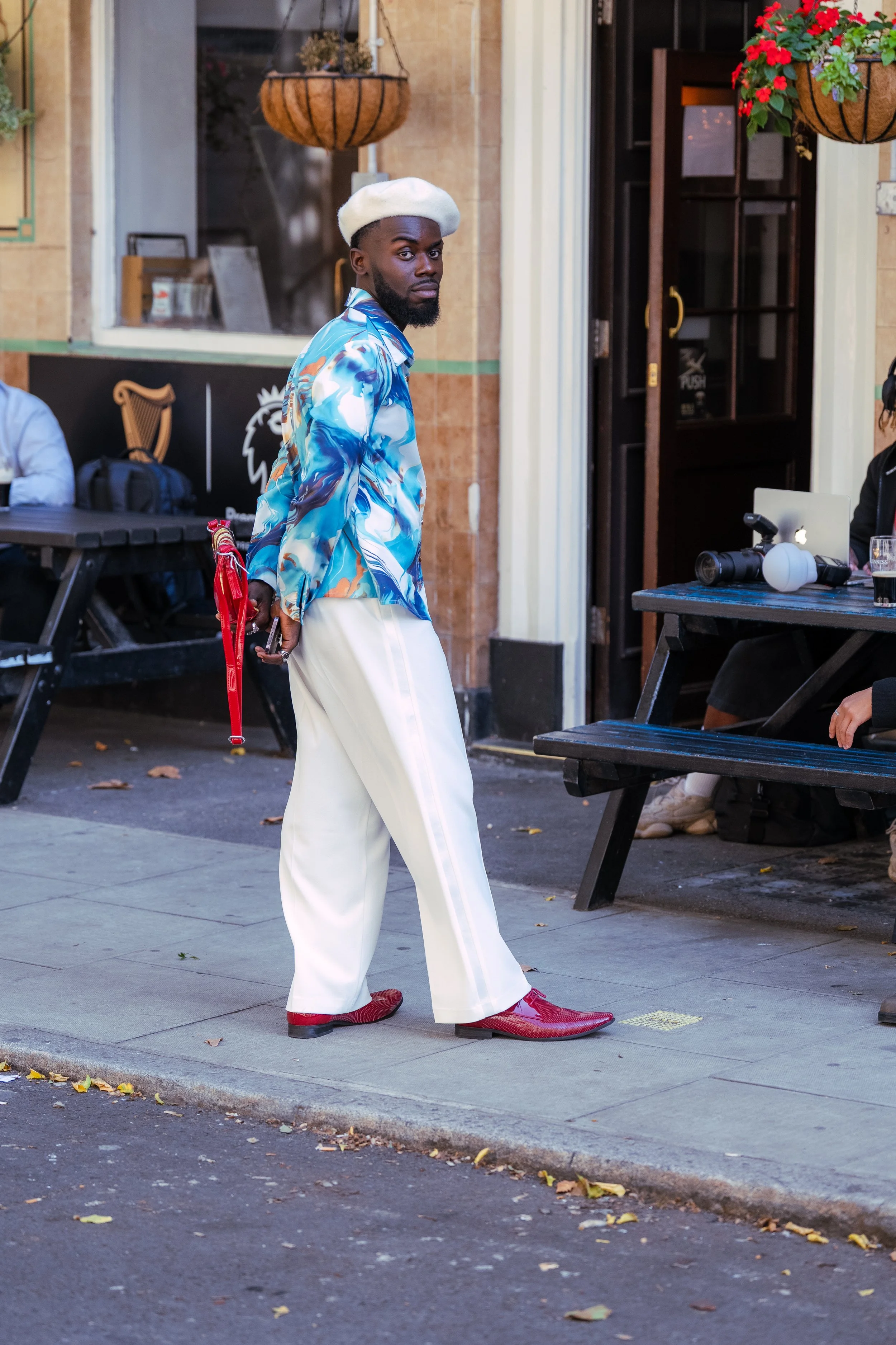 A man with a white beret, blue patterned shirt, white wide-leg trousers, and red shoes standing on a sidewalk outside a building with hanging flower baskets and outdoor seating.