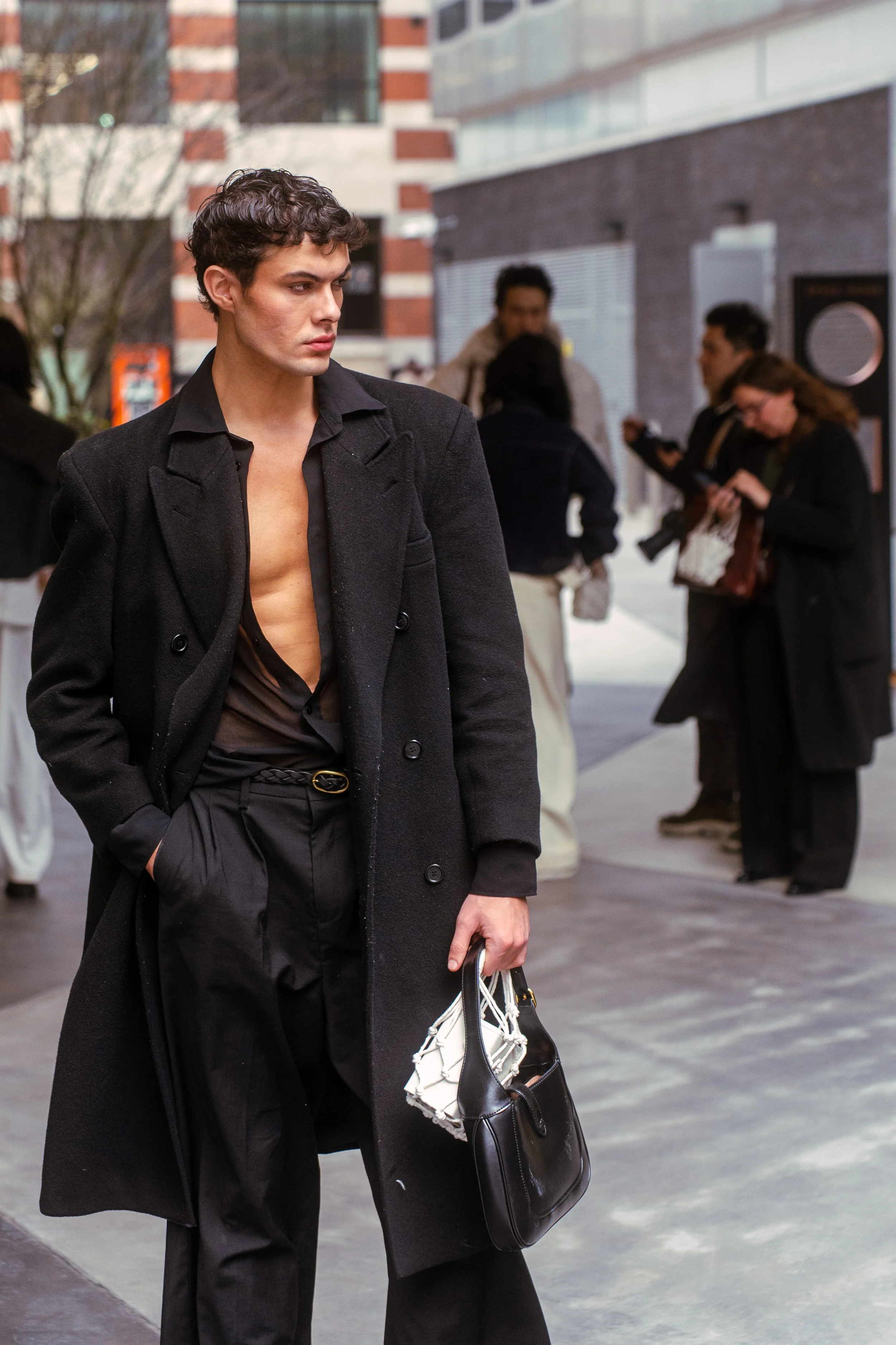 A young man with curly hair and fair skin dressed in black, wearing a long black overcoat, black pants, and a partially unbuttoned shirt, holding a black handbag, walking on a city sidewalk with other people in the background.