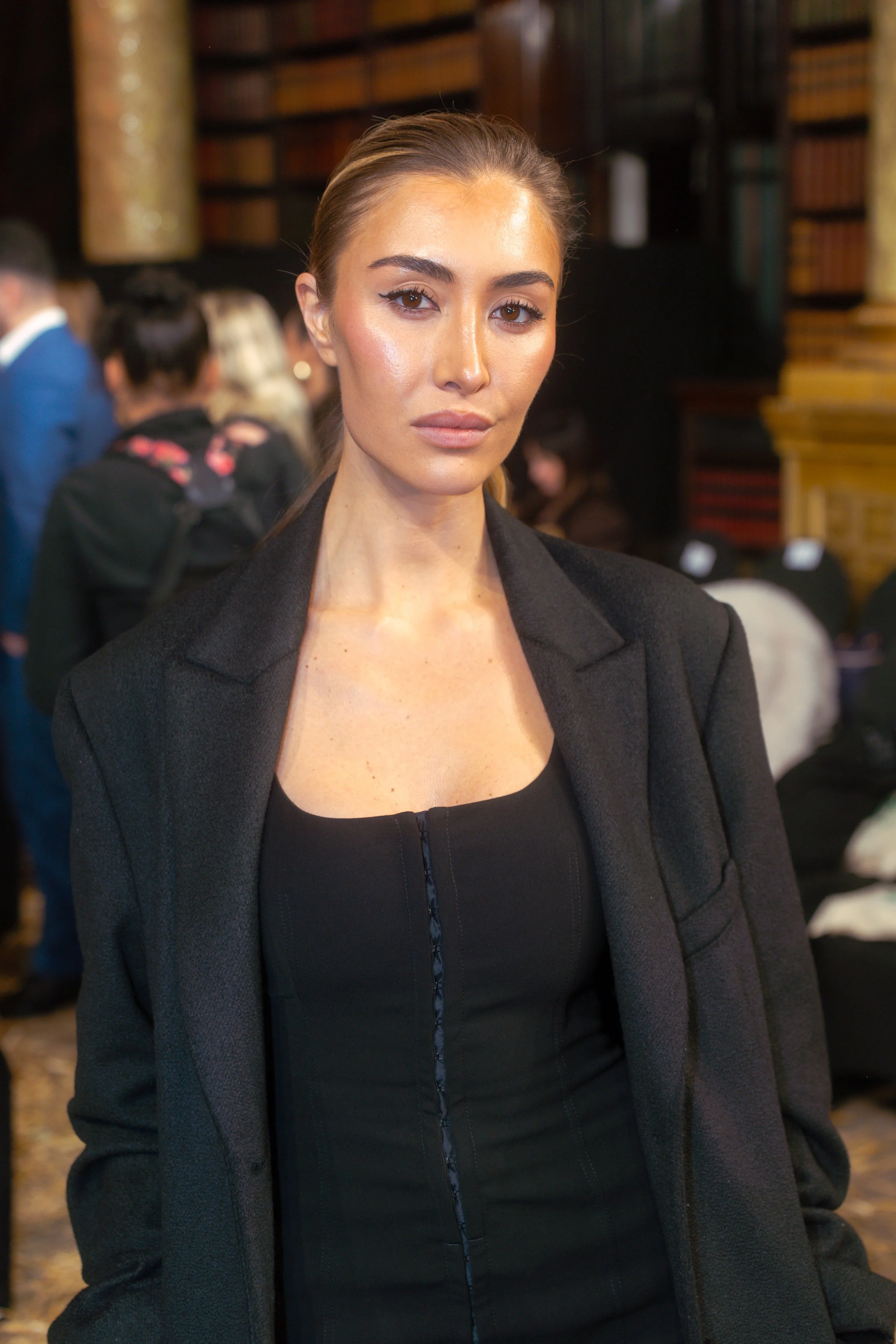 A woman with light skin, brown hair tied back, wearing a black top and black blazer, standing indoors with bookshelves and several people in the background.