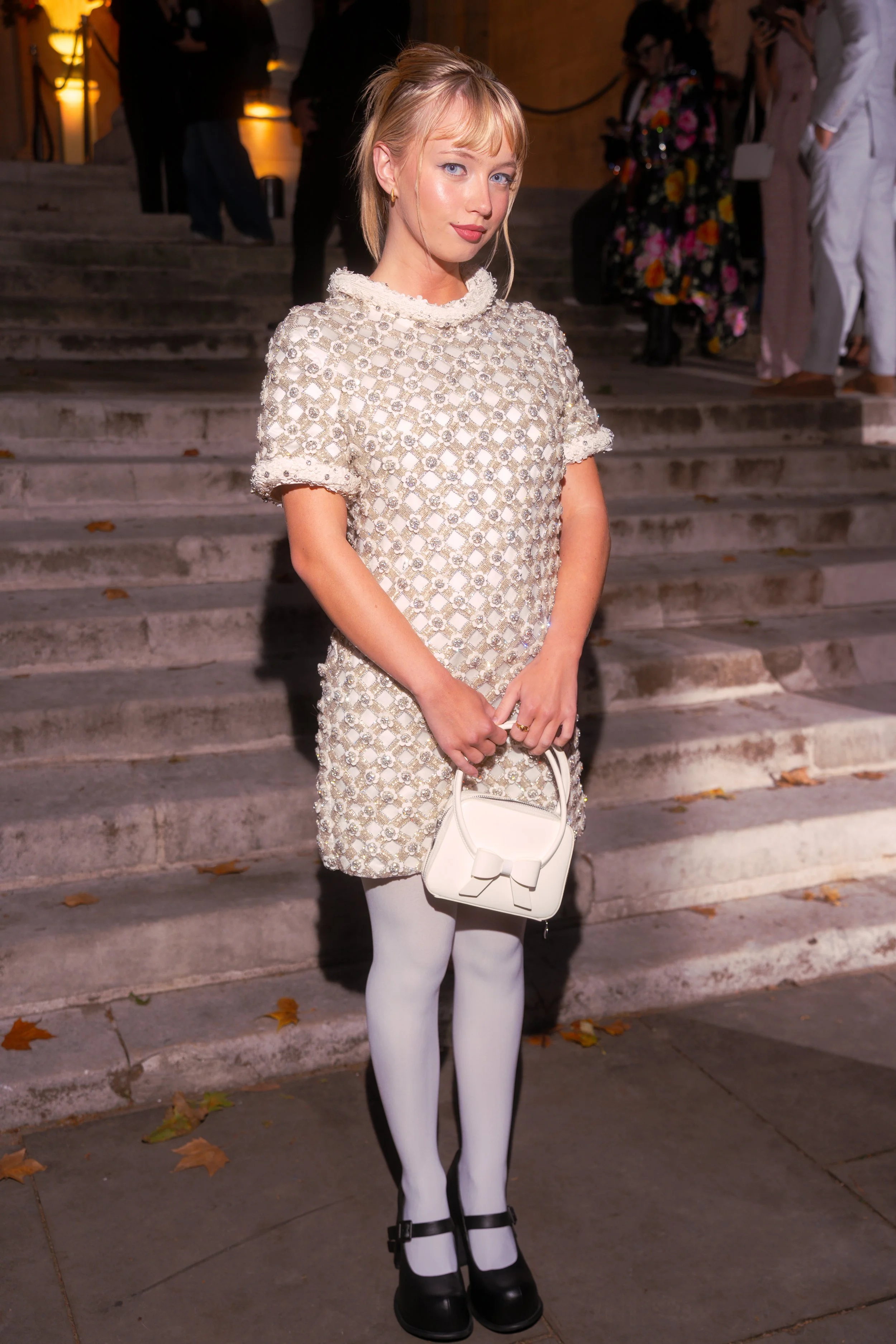 A young woman with blonde hair and blue eyes, wearing a pearl-embellished dress, white tights, black shoes, and holding a white handbag with a bow, standing on steps at an evening event.