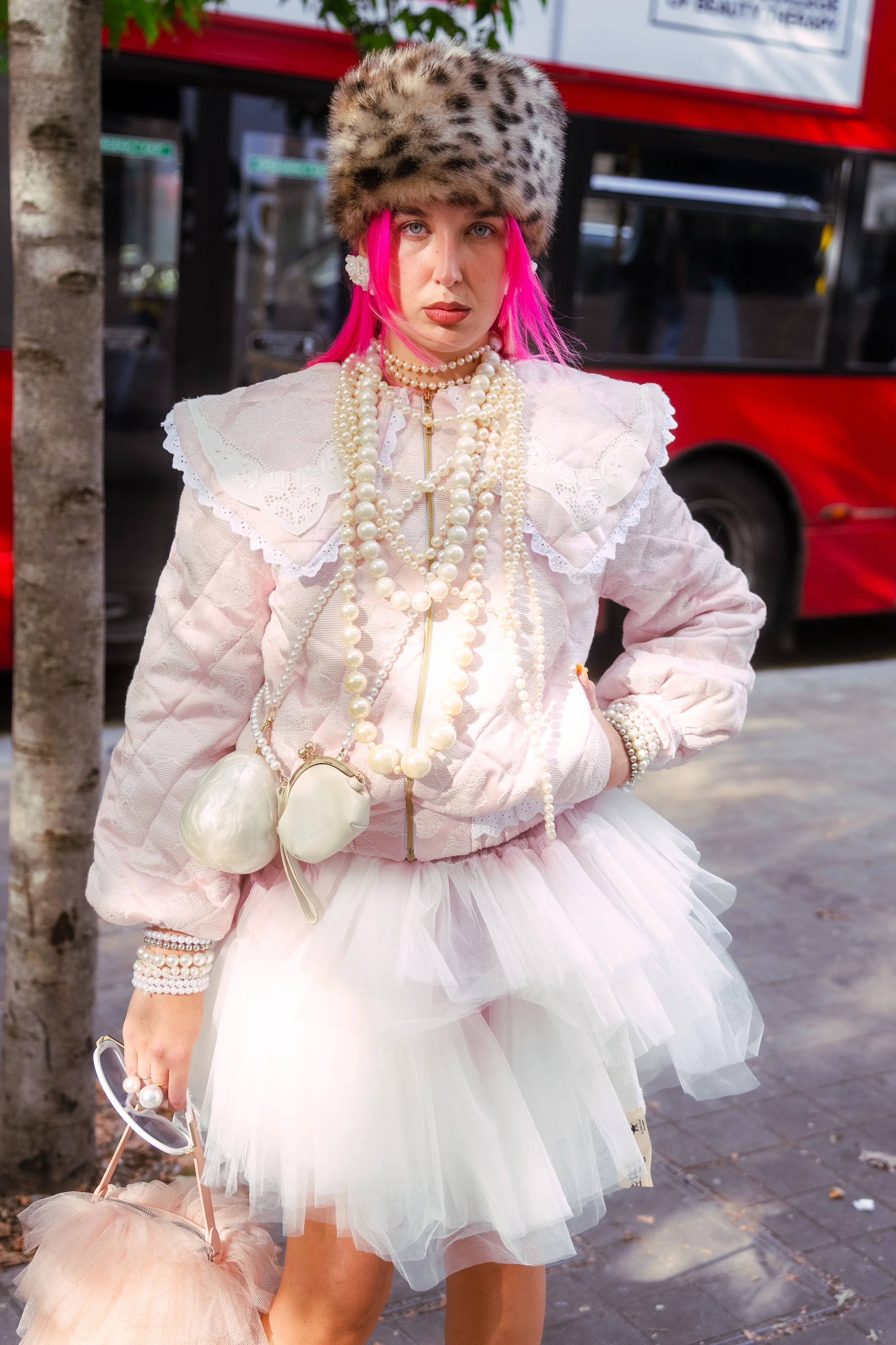 A woman with bright pink hair standing on a city sidewalk in front of a red bus. She is wearing a faux fur hat, a white jacket with lace details, a tulle skirt, and multiple pearl necklaces. She has a small purse and is holding a pastel-colored umbre