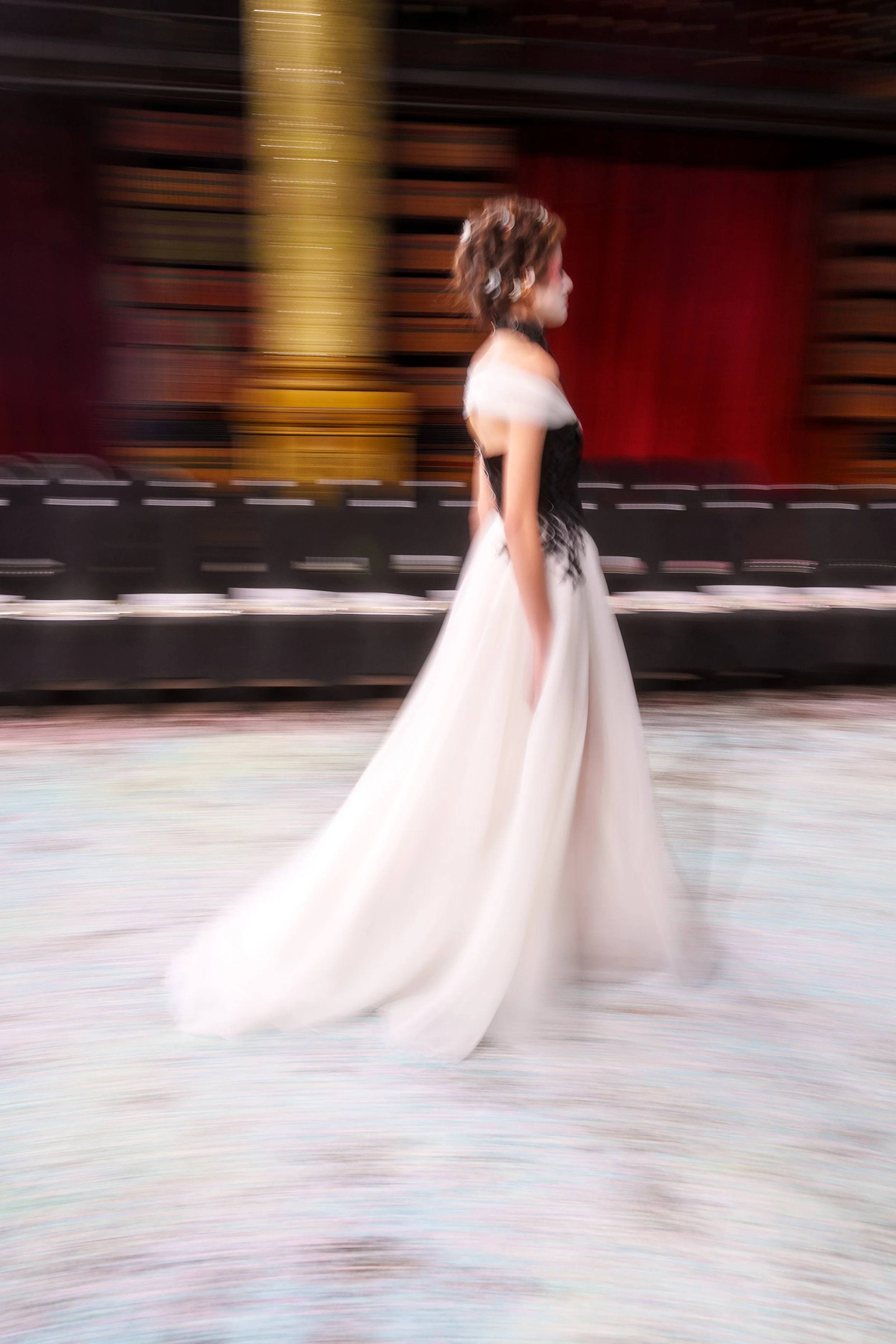 A woman wearing a black and white gown walking in a large, ornate hall with red curtains and gold accents, captured with motion blur.