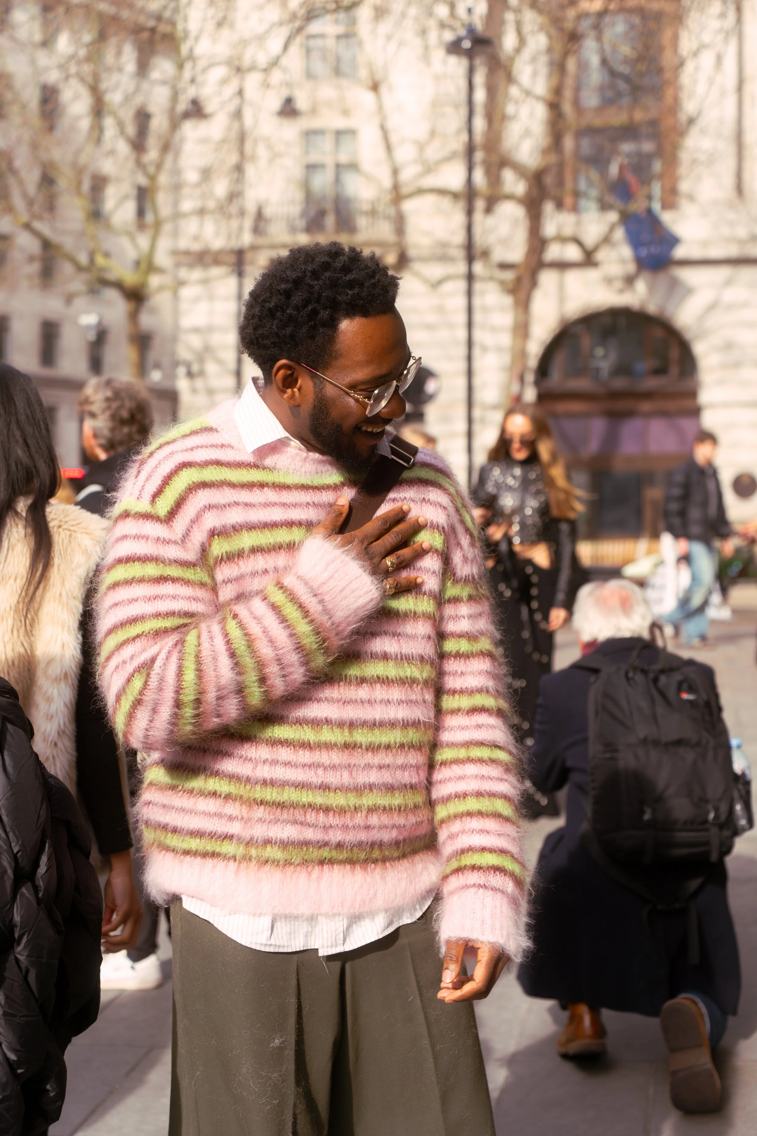 A man wearing glasses, a pink striped fuzzy sweater, and gray pants is outdoors on a sunny day, smiling and touching his chest, surrounded by people in a city setting.