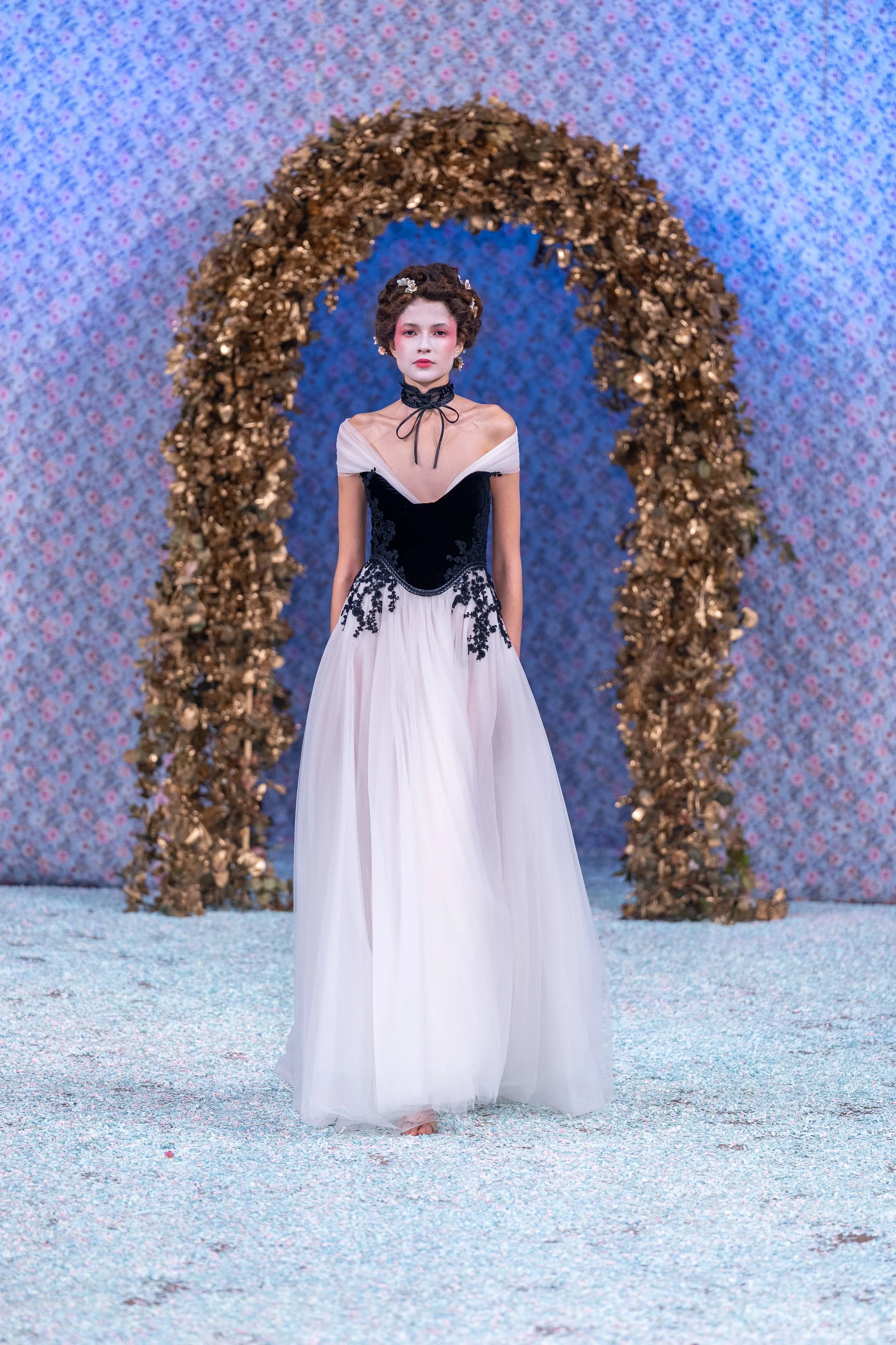 A woman in an elegant gown standing beneath a golden arch with a floral backdrop.