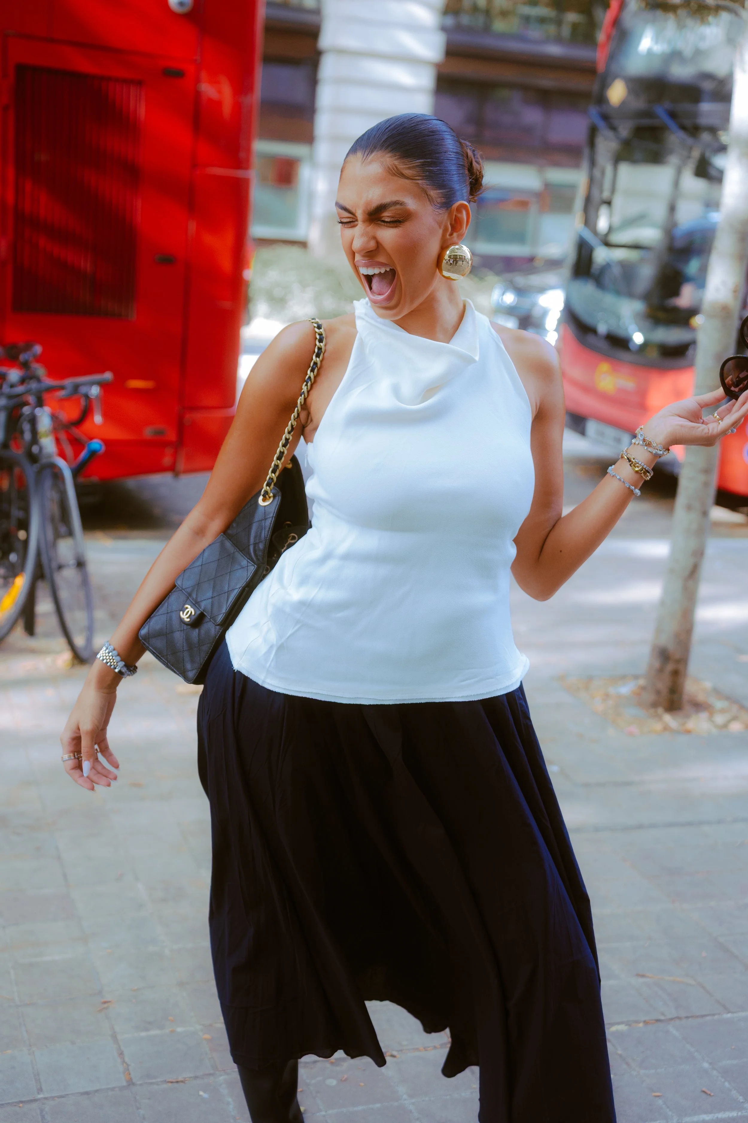 A woman with dark hair styled in a bun, wearing a white sleeveless top, black skirt, large gold earrings, and multiple bracelets. She is standing outdoors on a sidewalk with a broad smile, eyes closed, and appears to be laughing or shouting, with a l
