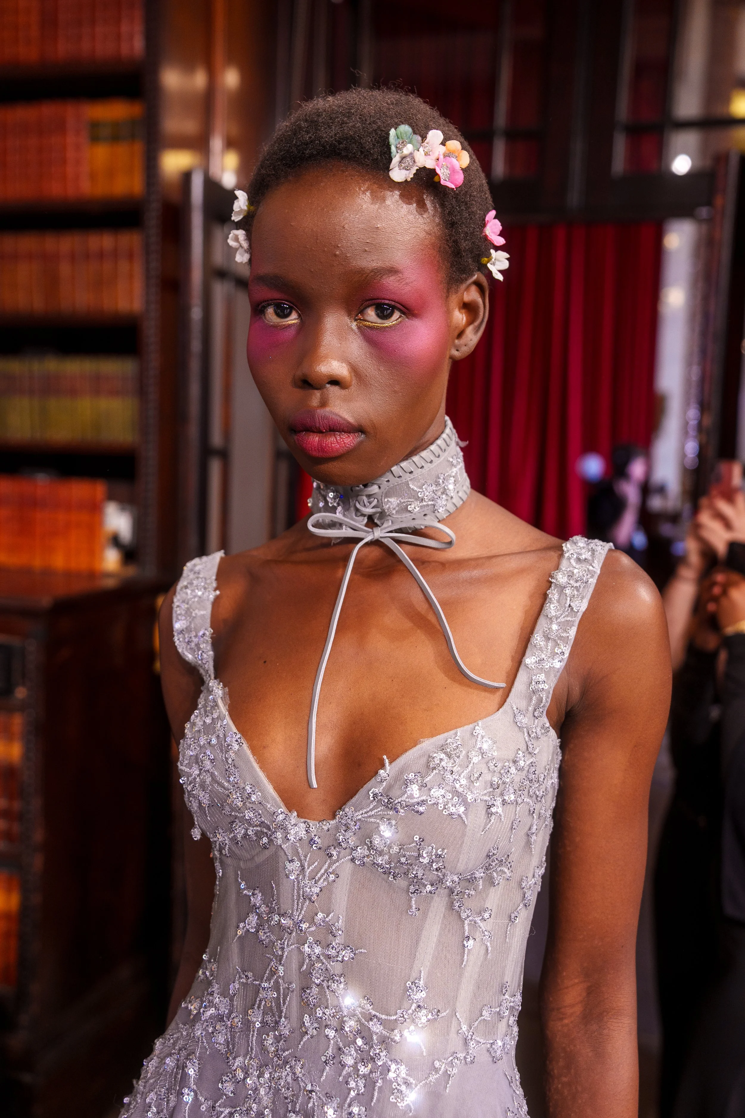 A young woman with dark skin and short hair styled with colorful hair accessories, wearing a detailed, glittery dress and a lace choker, standing indoors with a background of bookshelves and red curtains.